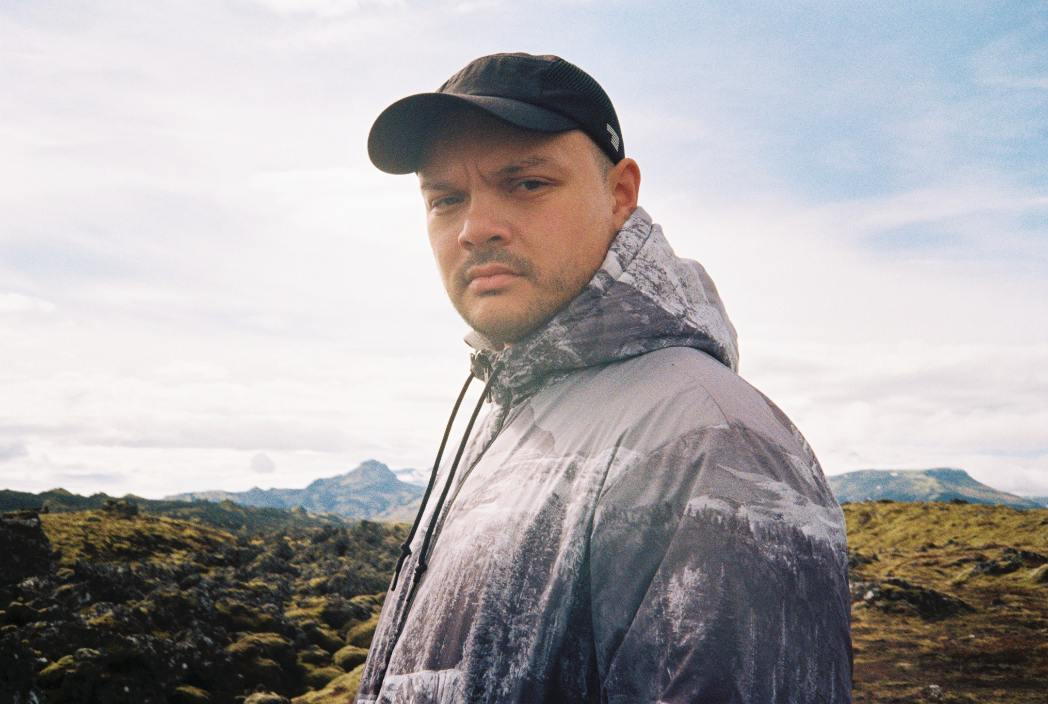 A man with a serious expression wearing a black cap and a patterned gray jacket standing outdoors in a mountainous landscape with a cloudy sky.