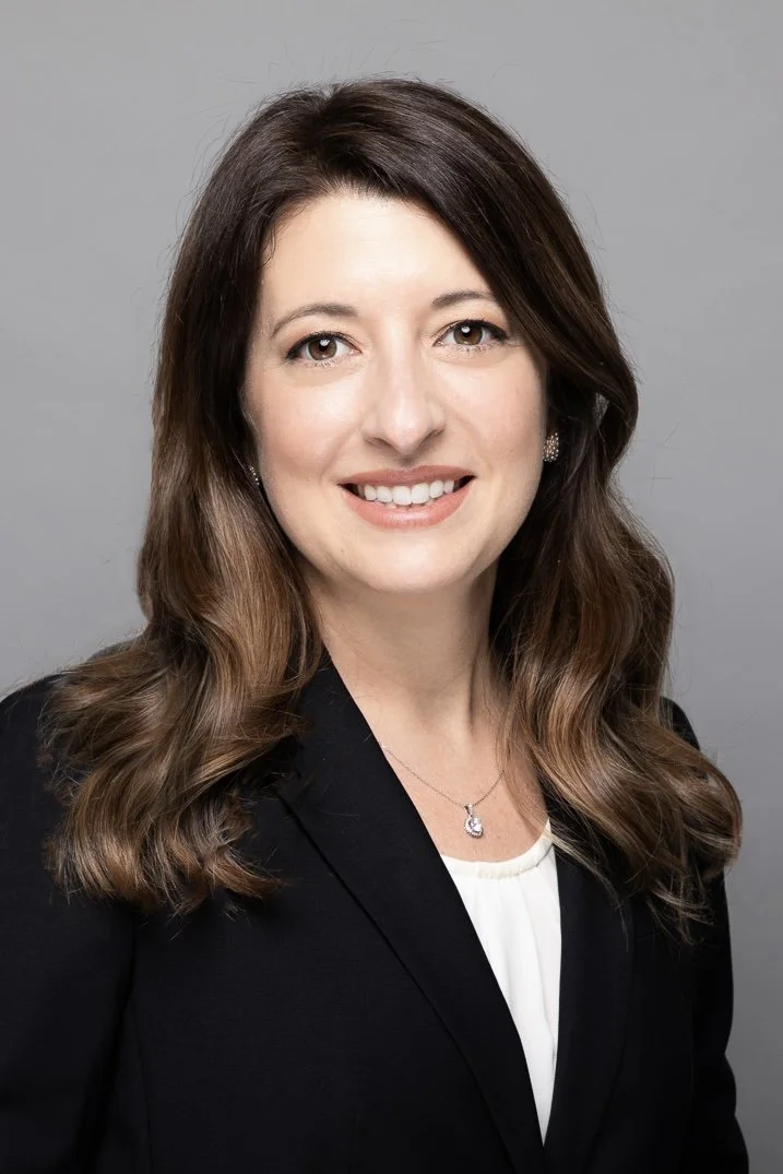 Professional headshot of a woman with long brown wavy hair, wearing a black blazer, white blouse, and jewelry, smiling against a gray background.