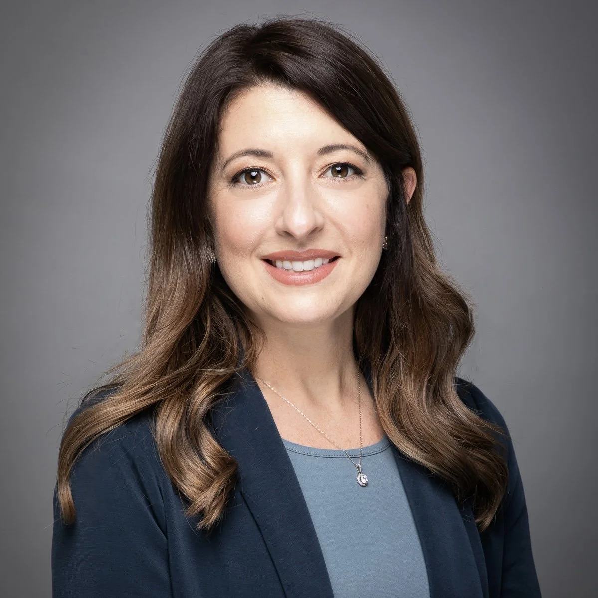 A professional headshot of a woman with shoulder-length brown hair, wearing a dark blazer over a blue top, smiling against a gray background.