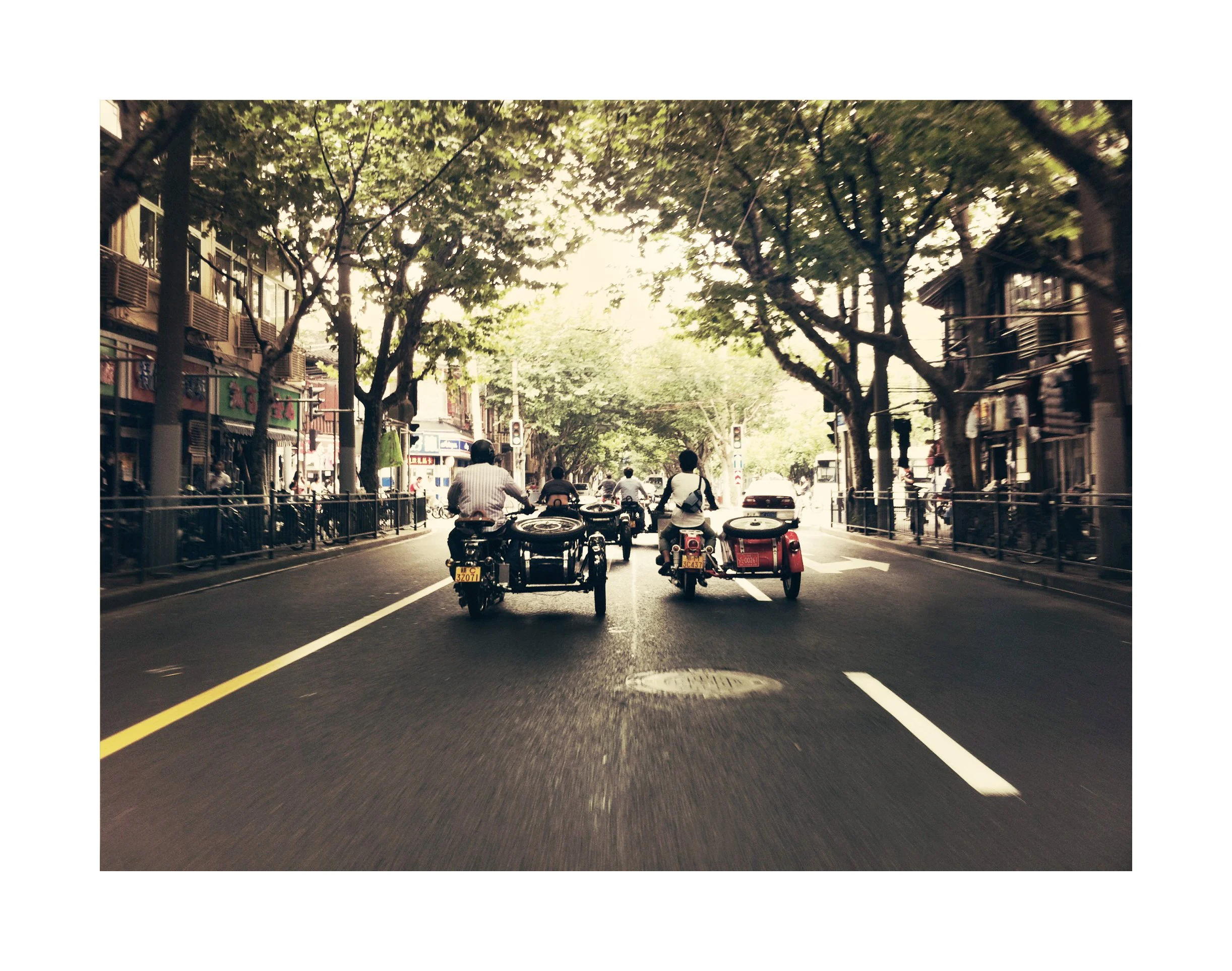 Rue urbaine avec trois motos et side-cars, arbres ligneux, bâtiments commerciaux et piétons en arrière-plan.