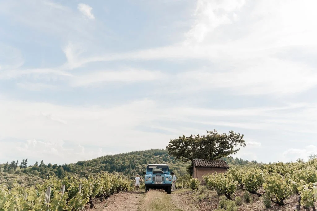 Vignoble du Beaujolais avec un chemin central, un petit bâtiment, un arbre, un véhicule ancien, et deux personnes dans un paysage champêtre sous un ciel partiellement nuageux.