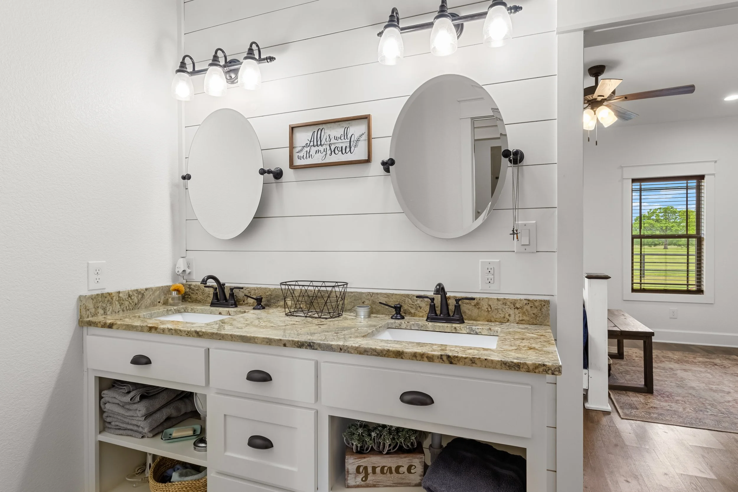 Bathroom vanity with dual sinks, granite countertop, and two round mirrors. White shiplap wall with a framed sign reading 'All is well with my soul'. There are black fixtures and a wire basket on the counter, with open shelves below storing towels, a
