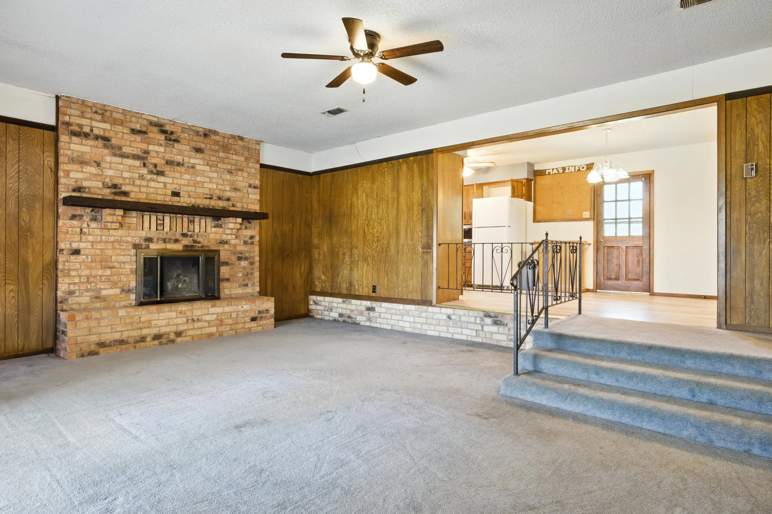 Living room with brick fireplace, wooden paneling, ceiling fan, and carpeted floor, opening to a dining area and kitchen with a refrigerator and a wooden door.