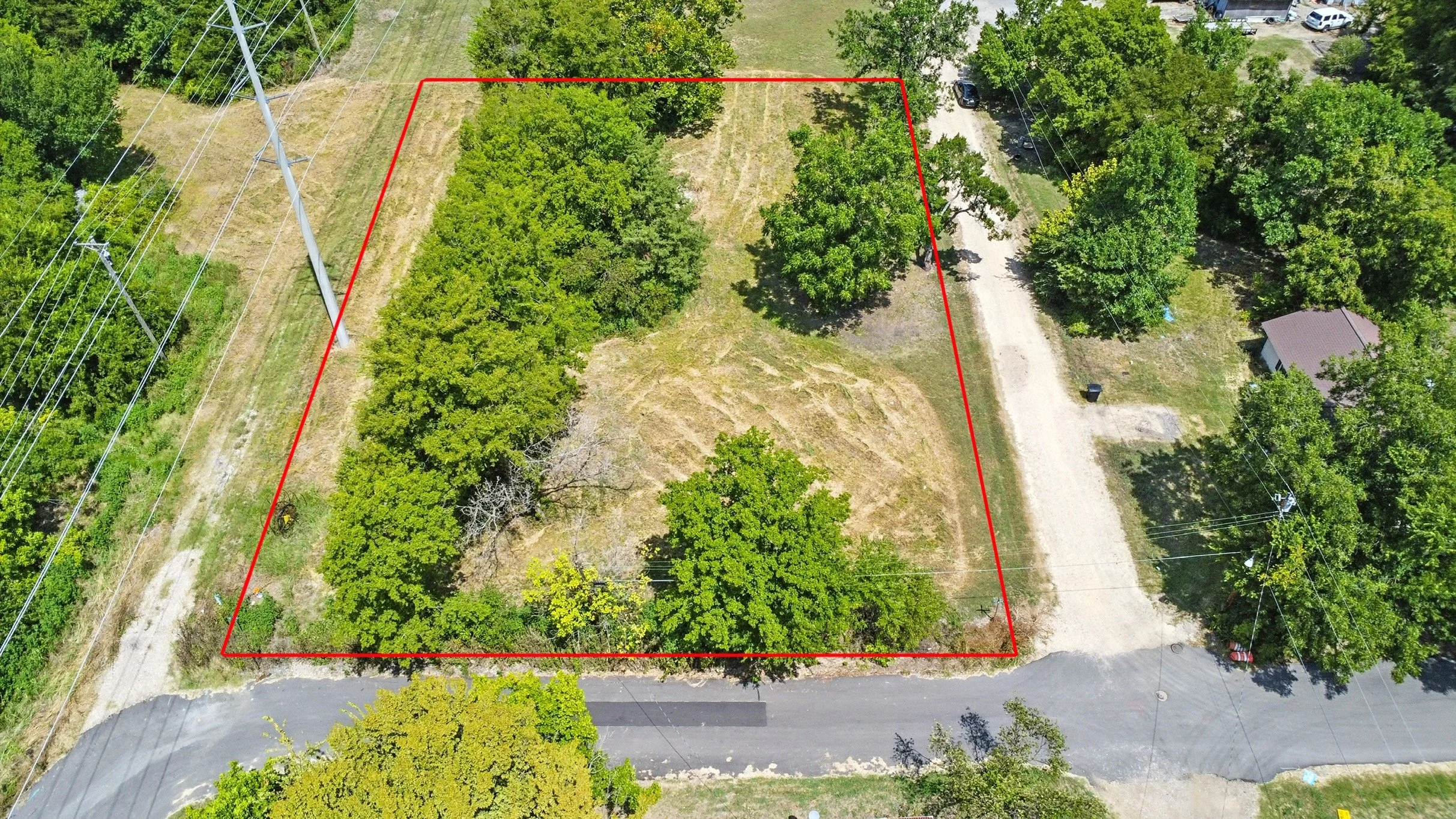 Aerial view of a vacant lot enclosed by red outline, with trees, grass, a gravel road on the right, and a paved road at the bottom, surrounded by residential and power lines.