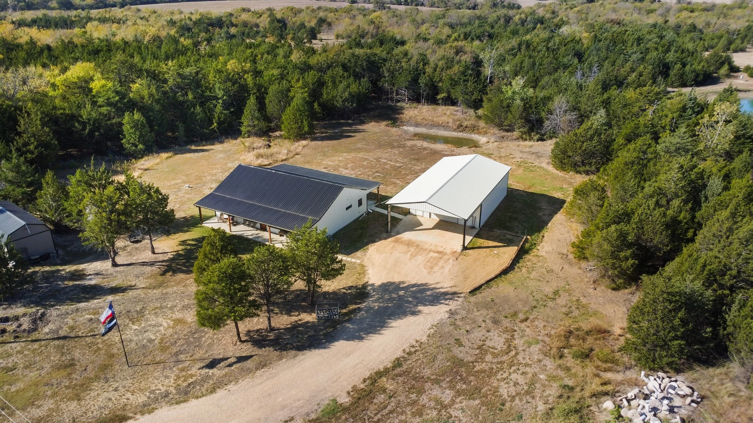 Aerial view of a rural property featuring two large metal buildings with white exteriors, one with a dark roof and the other with a light roof, surrounded by trees, dirt roads, and a grassy area with a few scattered trees, a small pond, and a flagpol