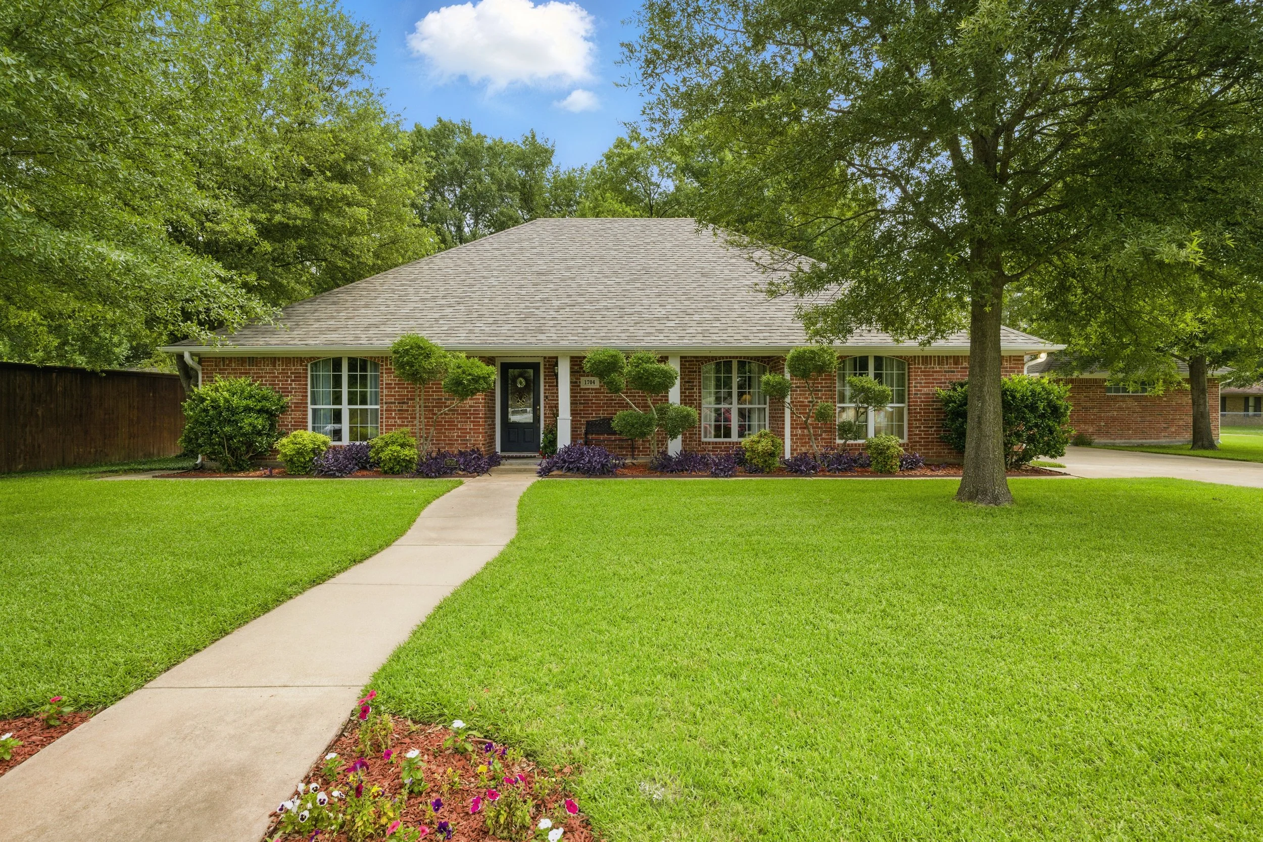 A single-story brick house with a gray shingle roof, large front windows, and a black front door with a wreath. The house is surrounded by a well-maintained green lawn, trees, shrubs, and flower beds, with a concrete walkway leading to the front door