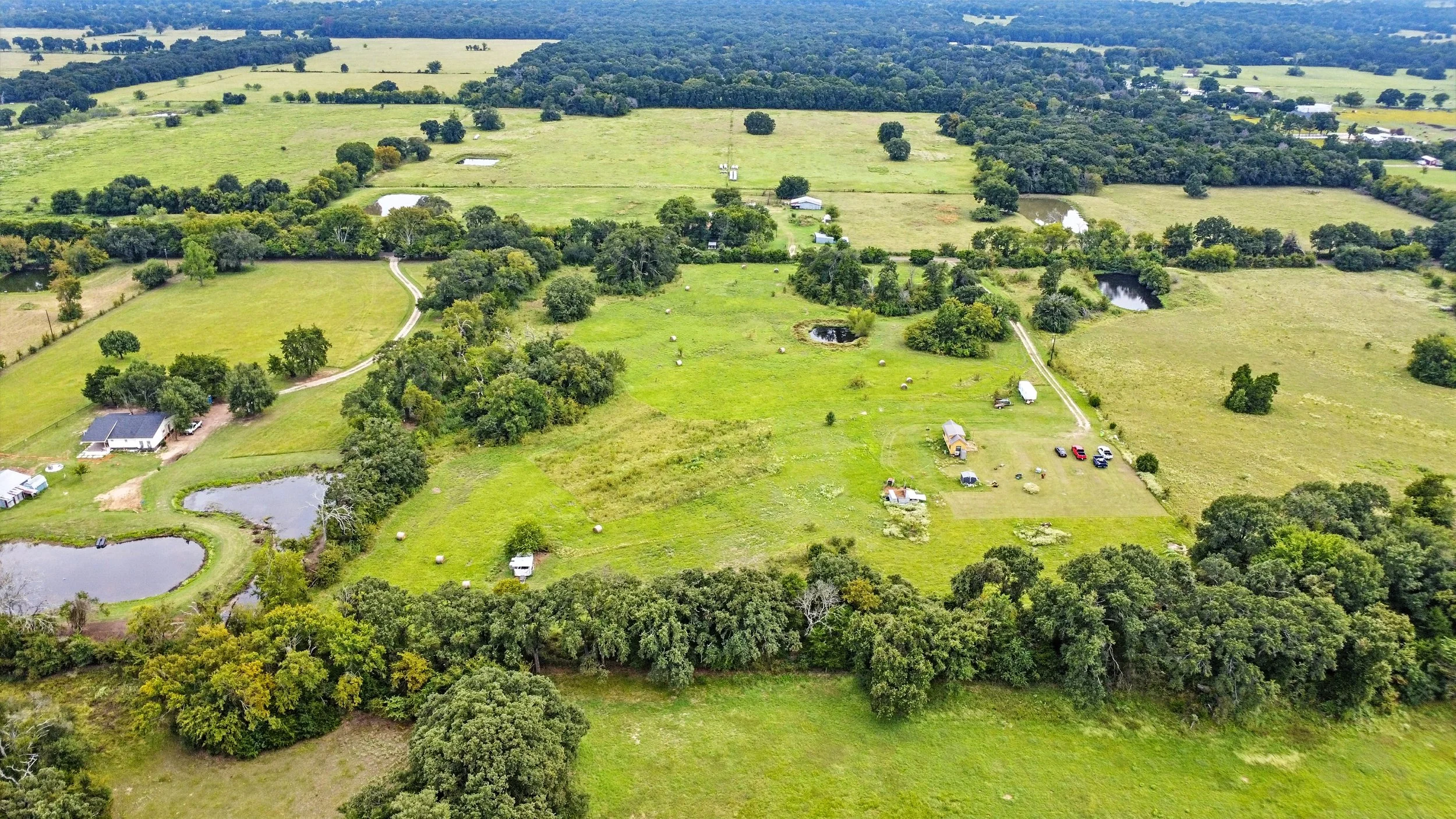 Aerial view of a rural landscape with green fields, scattered trees, small ponds, and a few houses and vehicles.