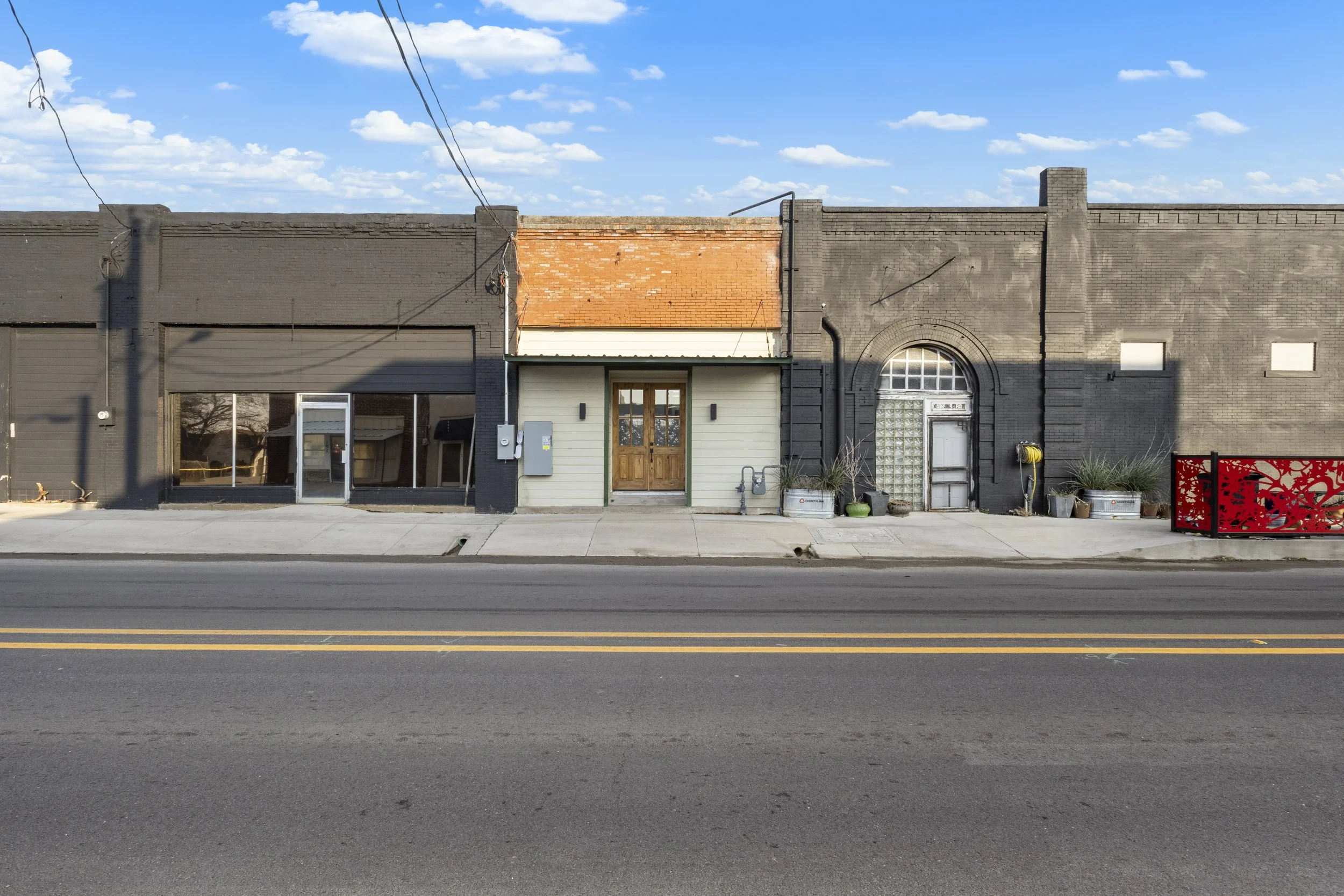 A row of small commercial buildings on a city street, with sidewalk in front and a blue sky with scattered clouds overhead.