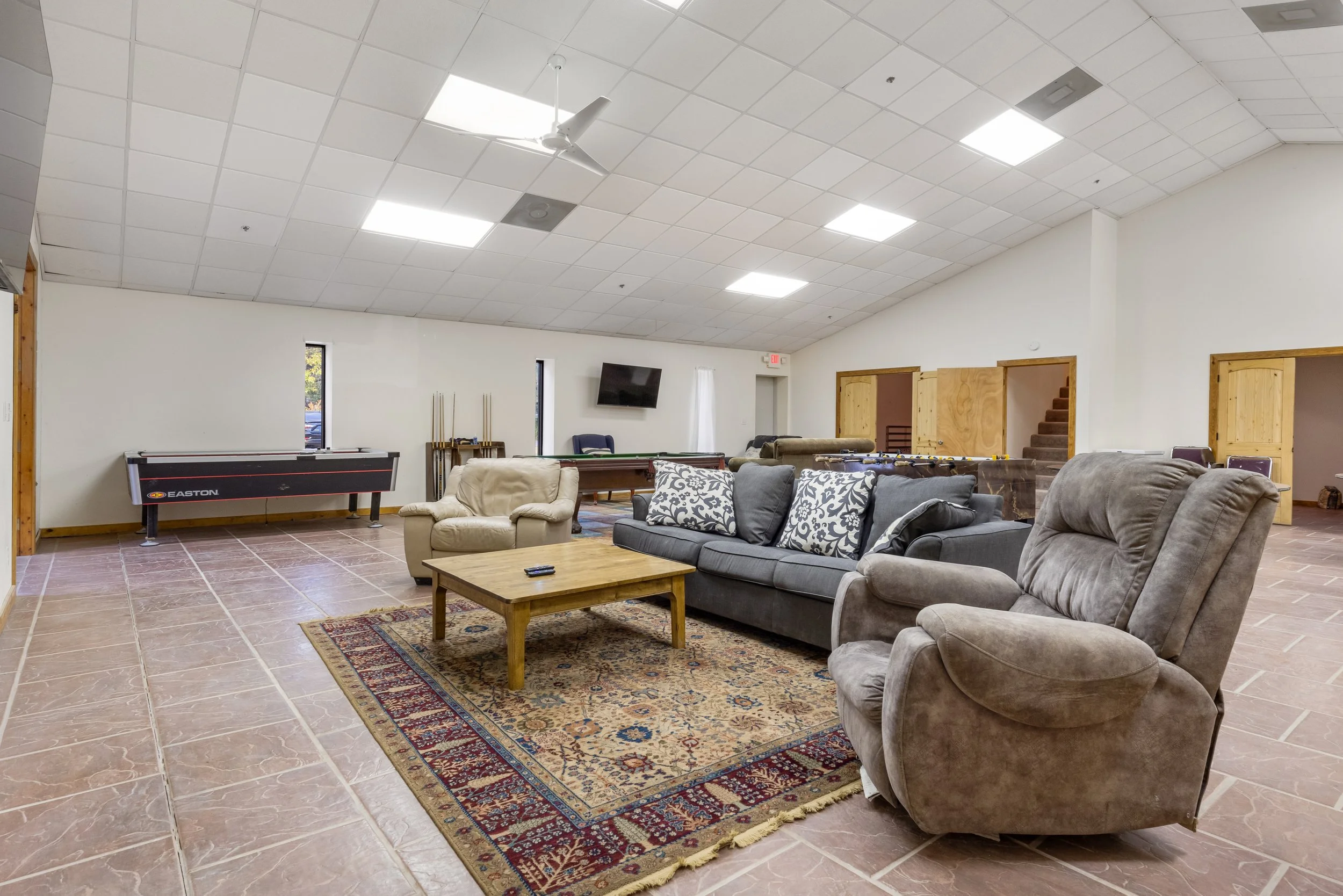 A spacious game room with tile flooring, featuring a sofa, armchairs, a coffee table, a pool table, an air hockey game, a flat-screen TV on the wall, and wooden doors leading to other rooms.