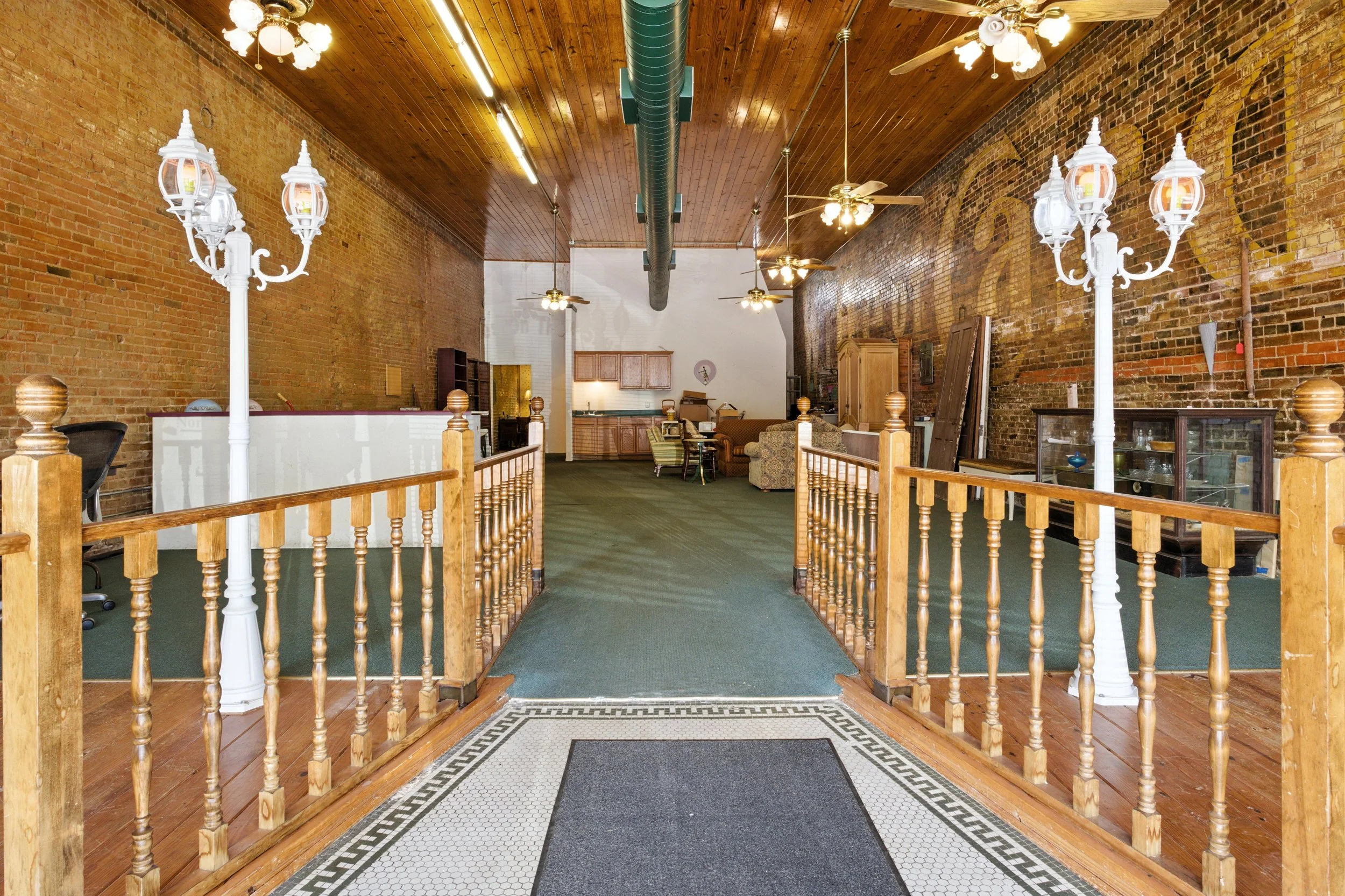 Interior of a vintage-style room with brick walls, wooden ceiling, green carpet, and white decorative lamps, with a small entrance area featuring a black doormat.