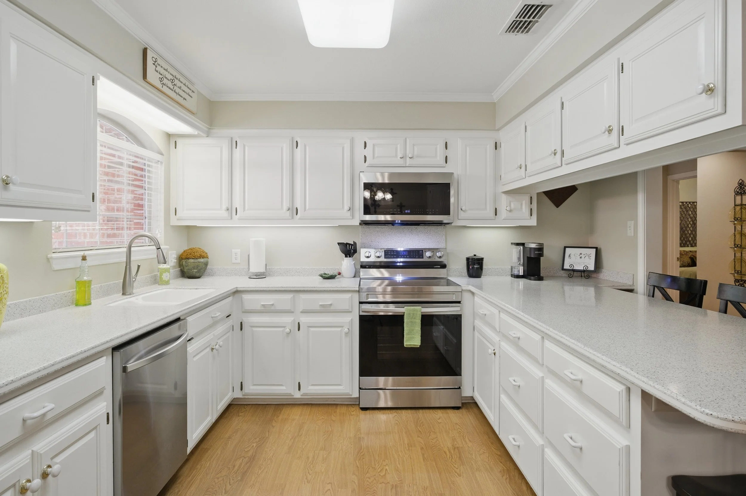 Kitchen with white cabinets, stainless steel stove and microwave, white countertop, window above the sink, and wooden flooring.