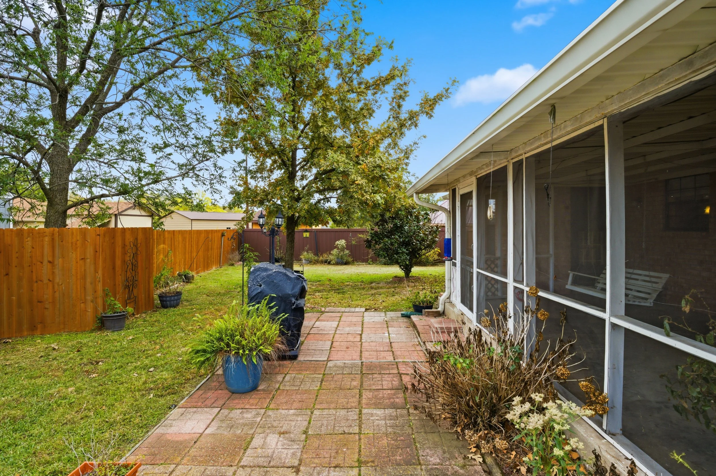 Backyard patio with brick paving, potted plants, trees, a wooden fence, and a screened porch with dried plants outside.
