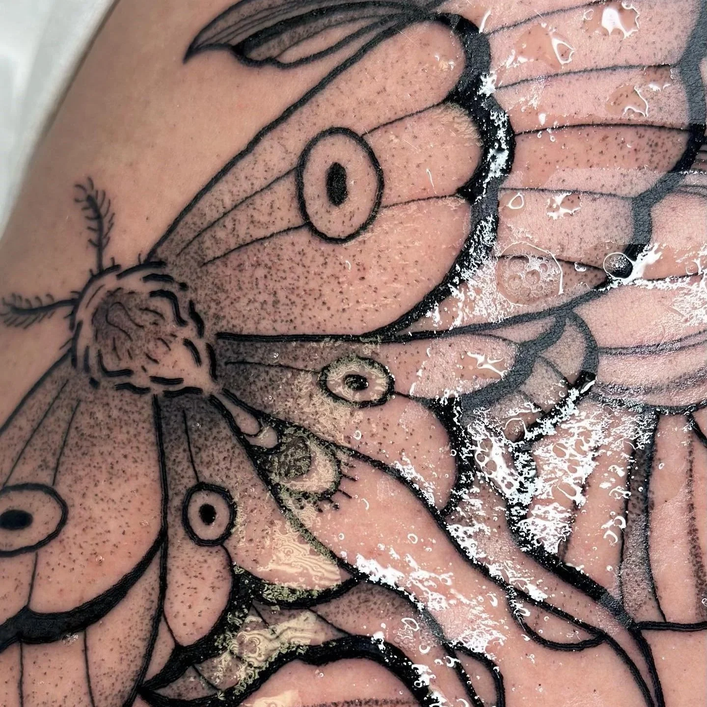 Close-up of a tattoo design of a face with black outlines, shading, and dotted details, with water droplets on the skin.