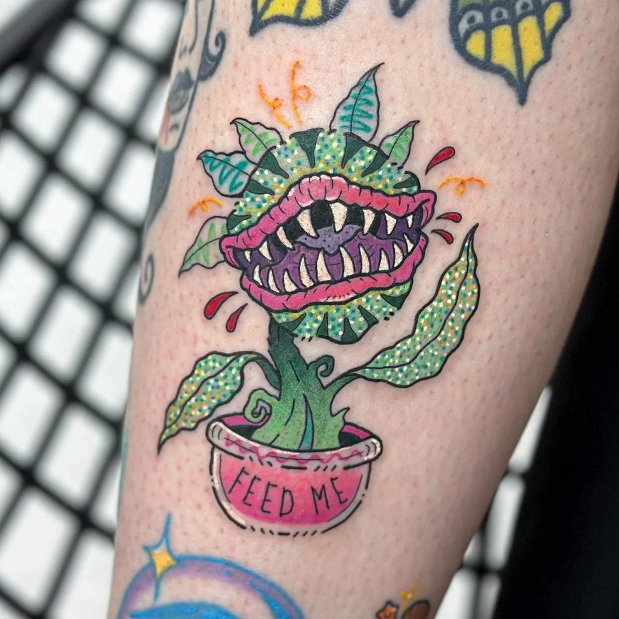 A colorful tattoo of a plant with a flower that has a wide open mouth with sharp teeth and a pink outer ring. The flower's stem is green in a pink pot labeled "FEED ME".