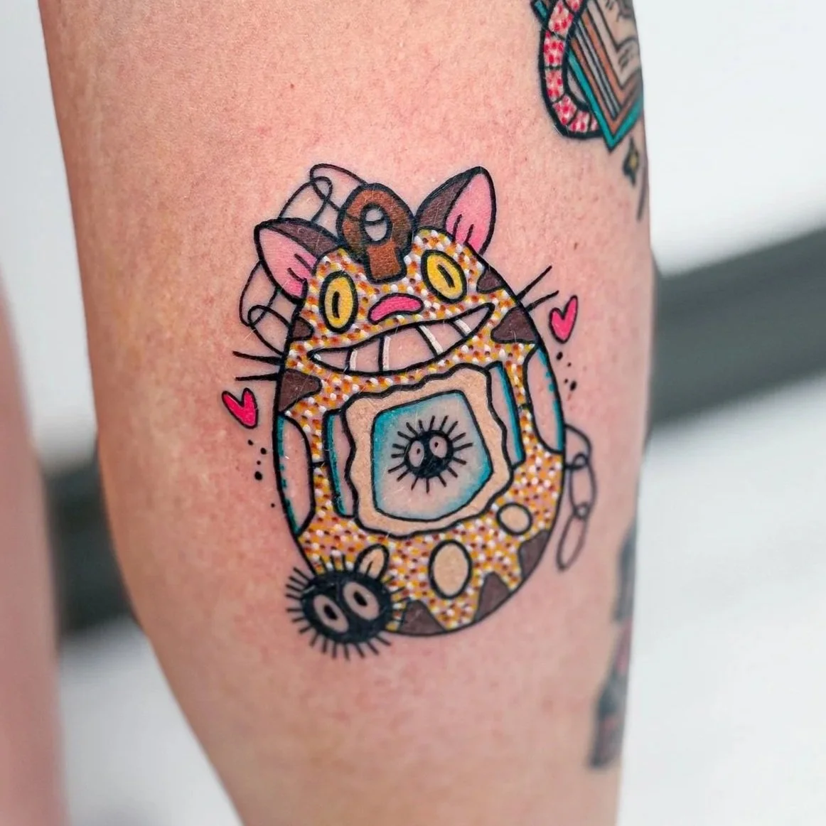 Colorful tattoo of a smiling cat with large yellow eyes, surrounded by hearts and abstract decorative elements.