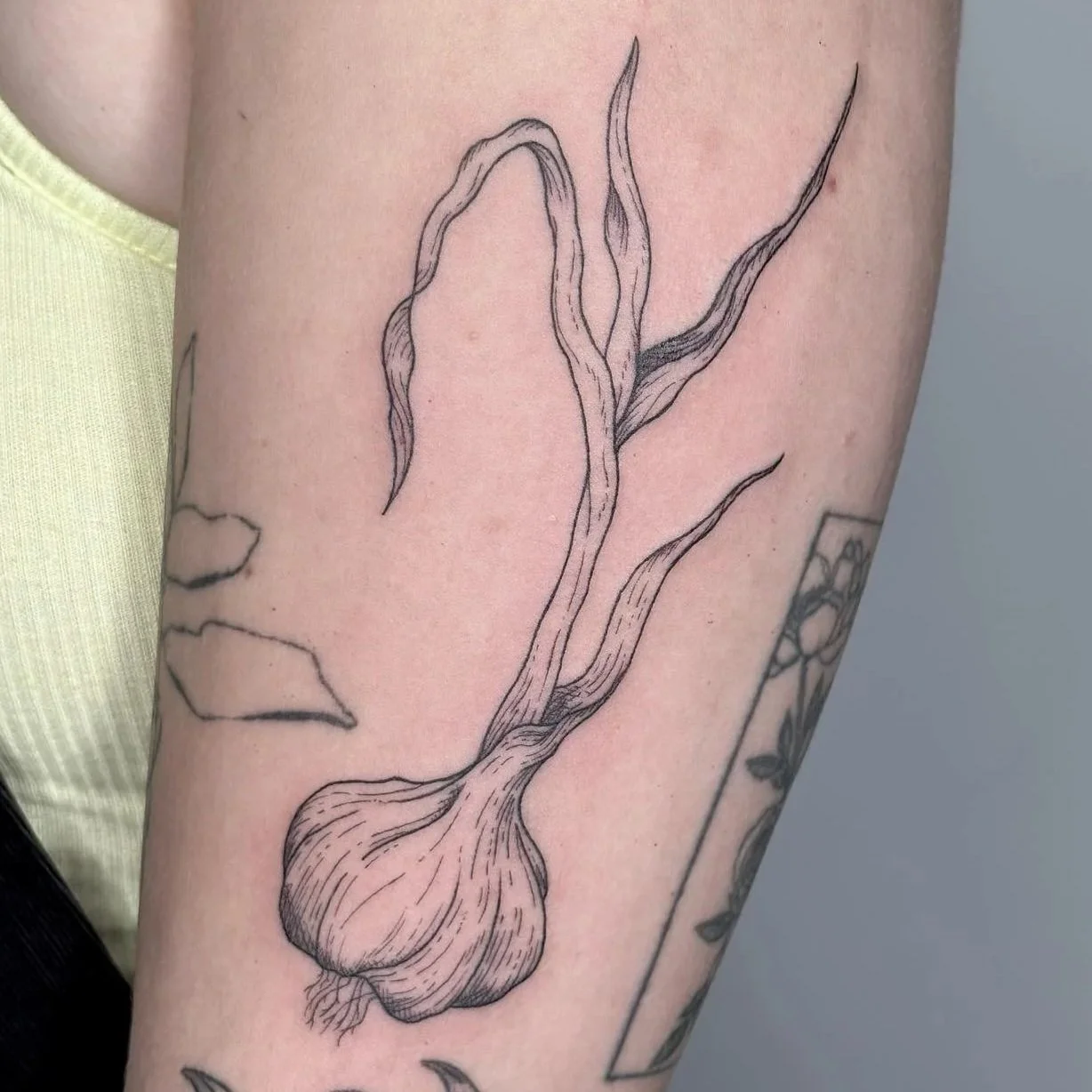 A black and gray line art tattoo of a sprouting garlic bulb on a person's upper arm.