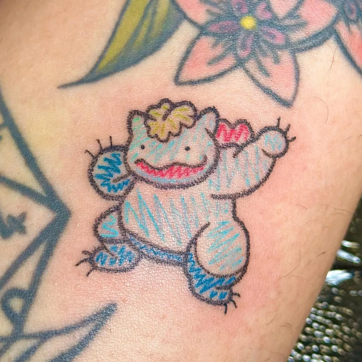 Colorful tattoo of a smiling cartoon frog with a flower on its head, waving, on skin with other tattoos visible in the background.