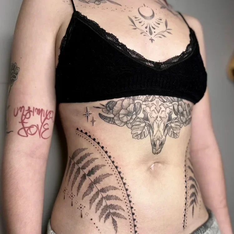 Close-up of a woman's torso with various tattoos, including a floral skull and moon design on her chest, fern leaf and dotted line tattoos on her stomach, and a red script tattoo on her upper arm, wearing a black lace crop top.