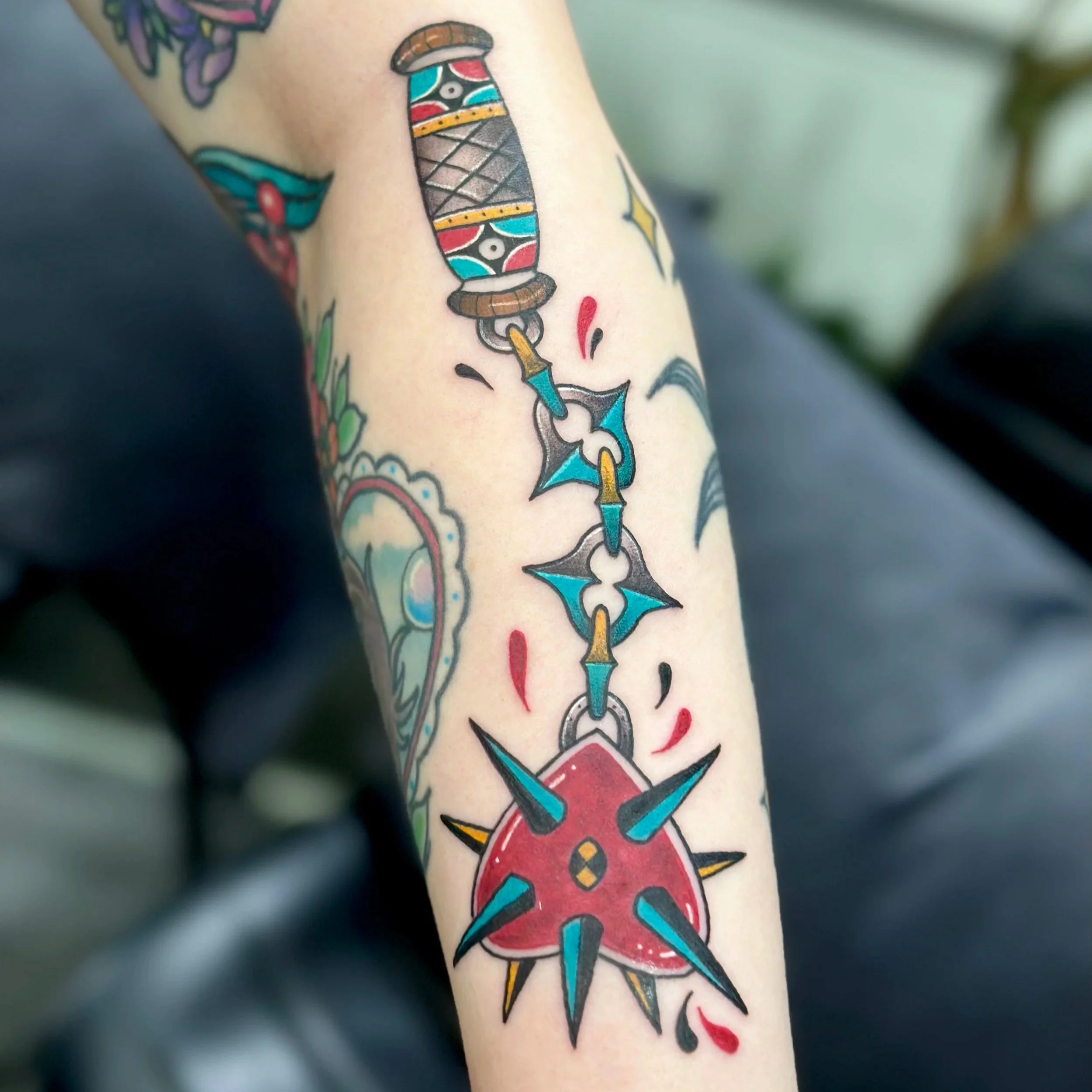 Colorful tattoo of a shuriken (throwing star) connected to a chain with a stylized, spiked red heart at the end on a person's arm.