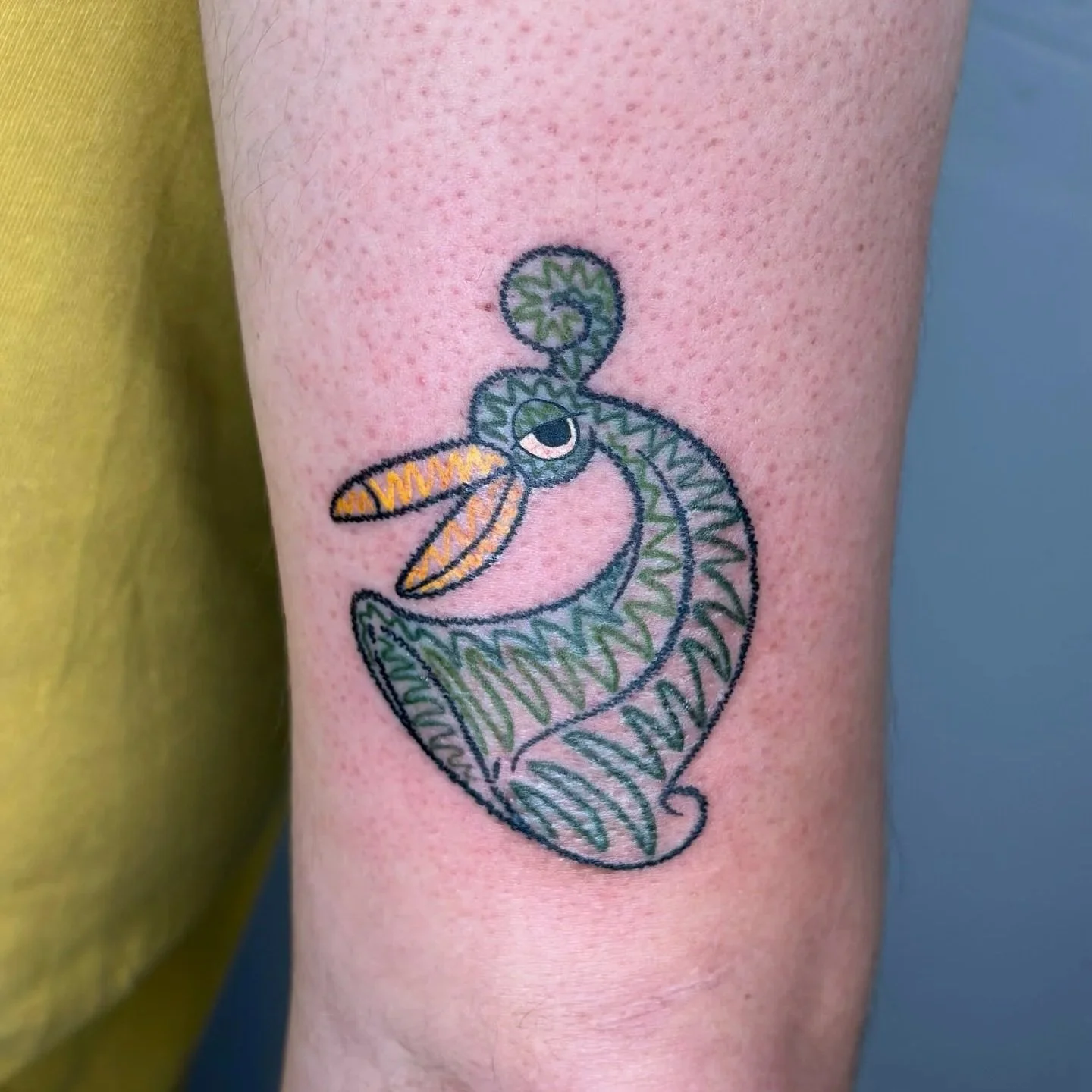 Tattoo of a stylized chameleon with a curled tail, green body, orange tongue, and black outlines on a person's arm.