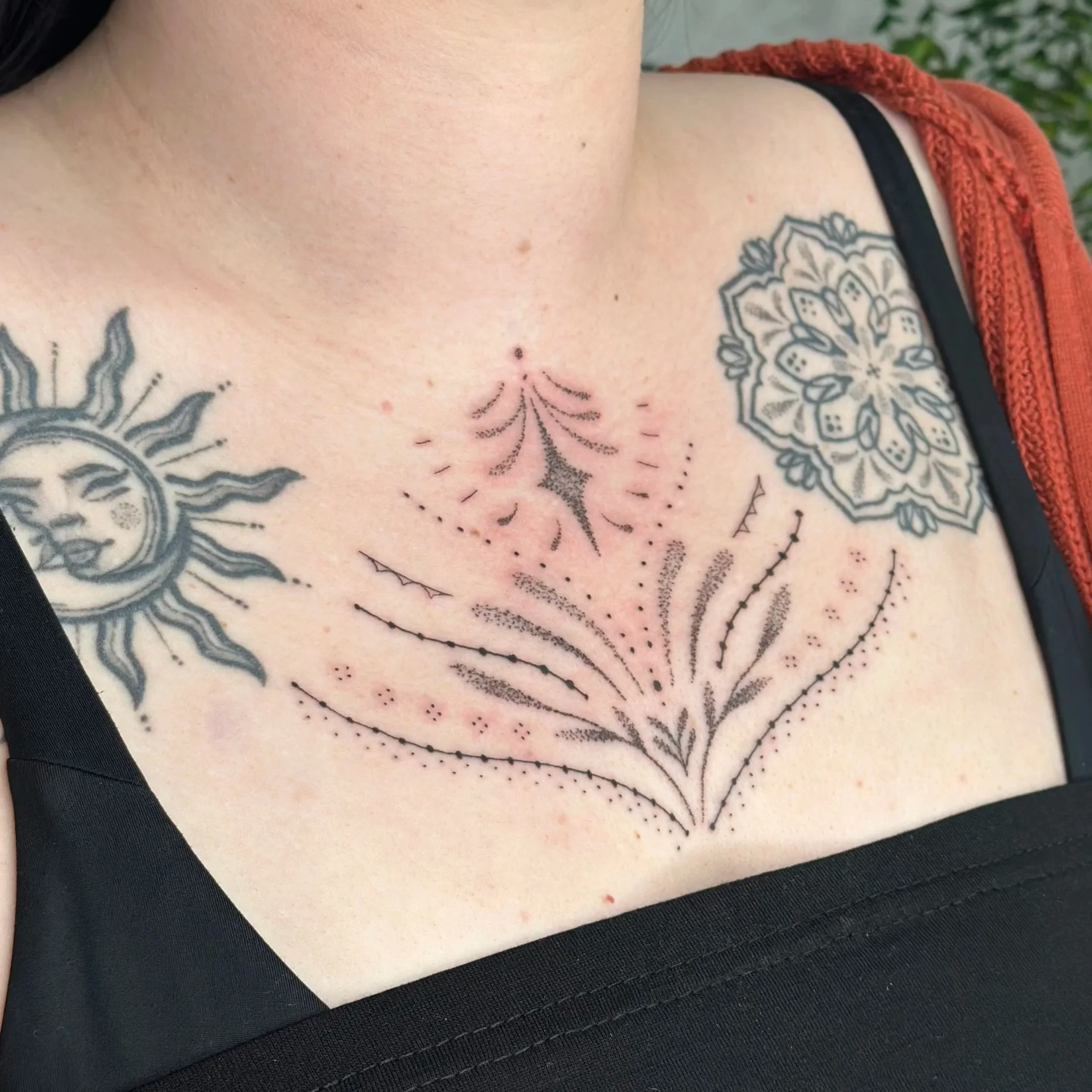 Tattoo of a stylized floral design with dotted lines, abstract shapes, and decorative elements on a person's chest, alongside additional tattoos of a sun with a face and a symmetrical floral pattern.