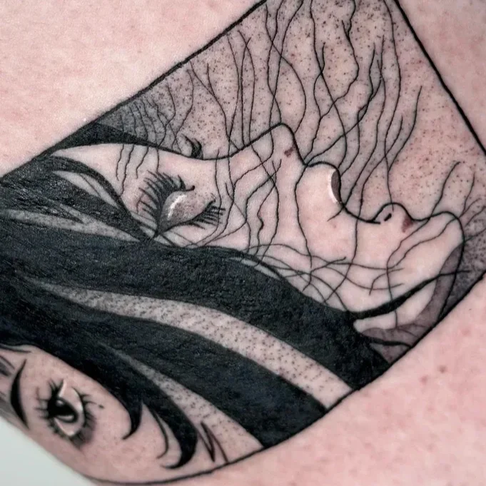 Close-up black and gray tattoo of a woman's face with long hair, with an emphasis on her eye, nose, and lips, on skin with some freckles.