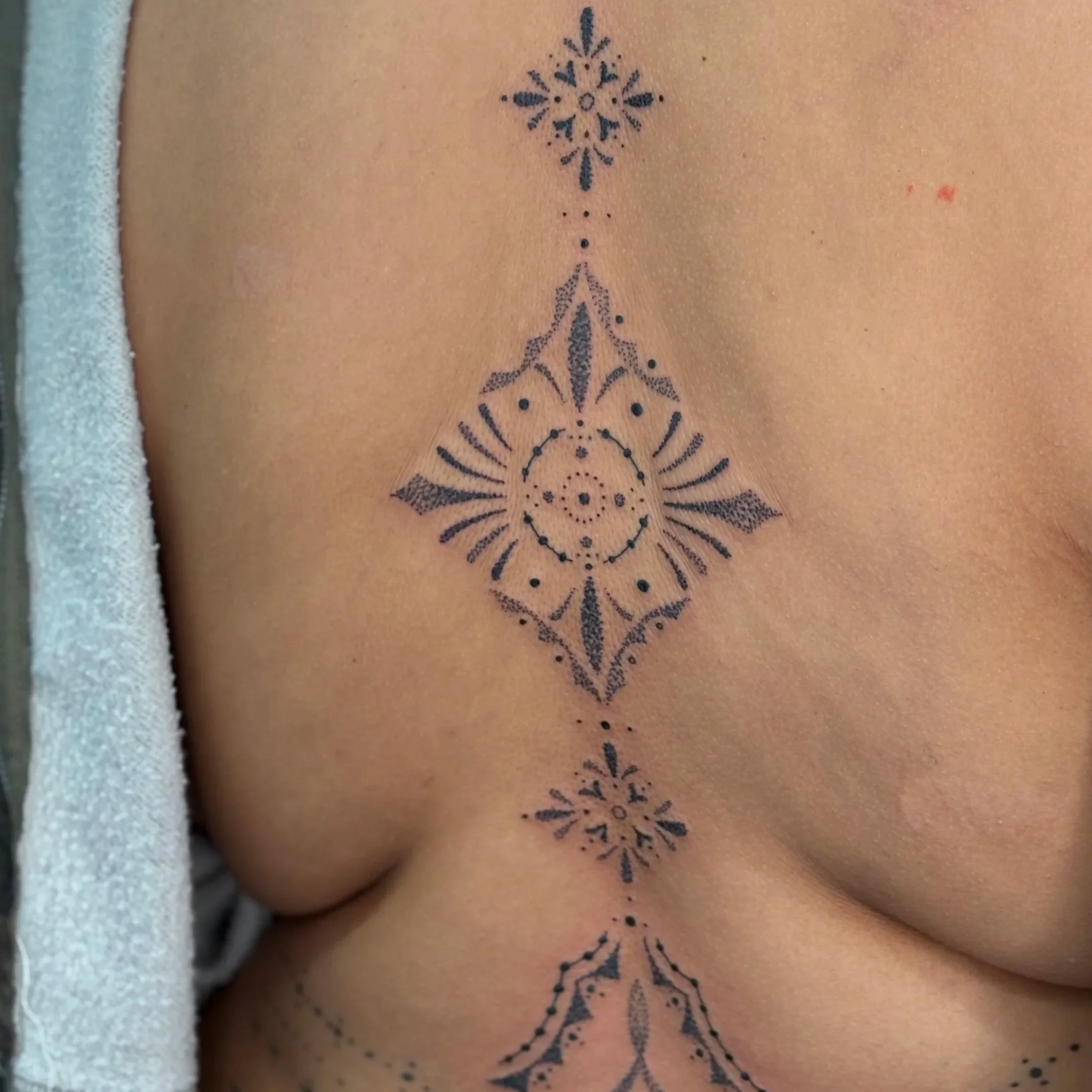 Close-up of a person's upper back featuring a symmetrical black tattoo with intricate geometric and floral patterns.