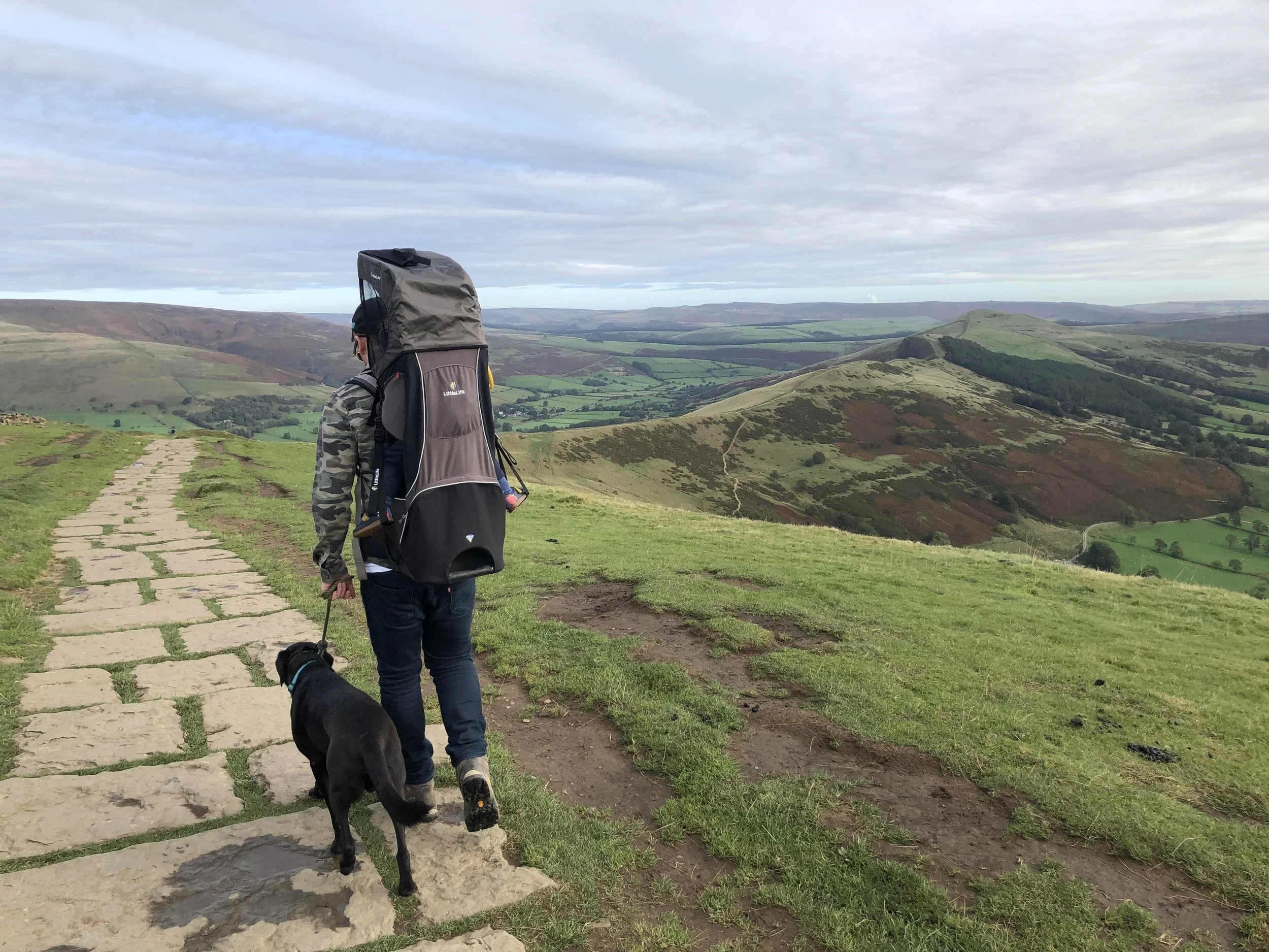 Person walking on a stone path with a large backpack, accompanied by a black dog, overlooking rolling green hills and valleys under a cloudy sky.