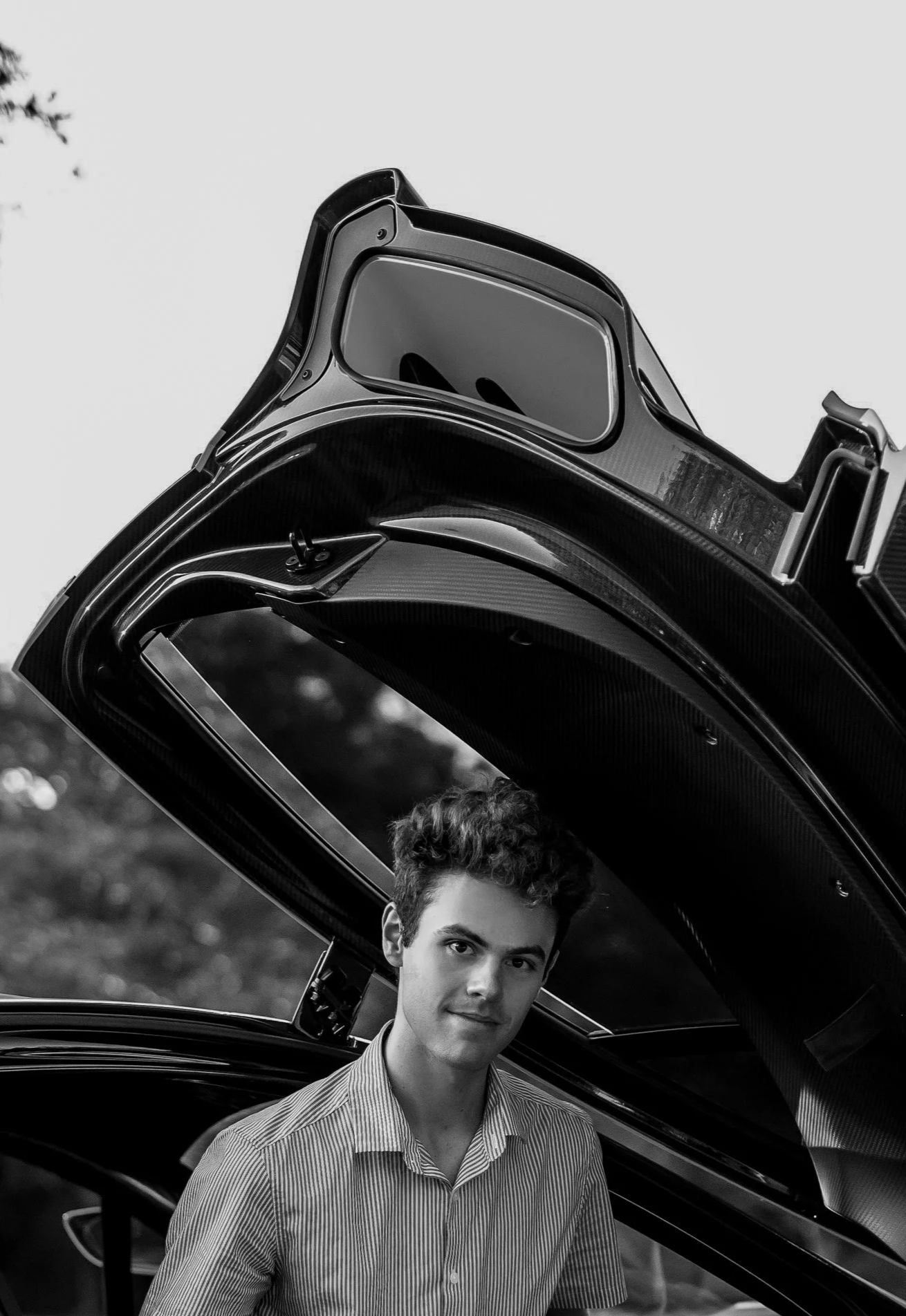 Young man with curly hair standing next to a sports car with its gull-wing door open, outdoors.