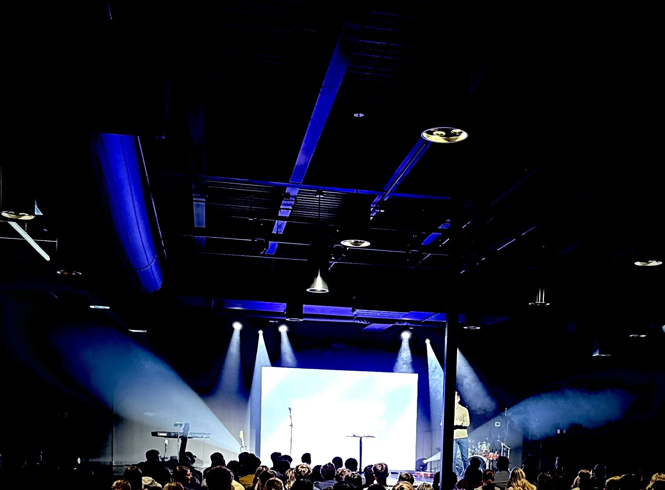Stage with blue and white lighting, audience watching a speaker, screen, and musical instruments.