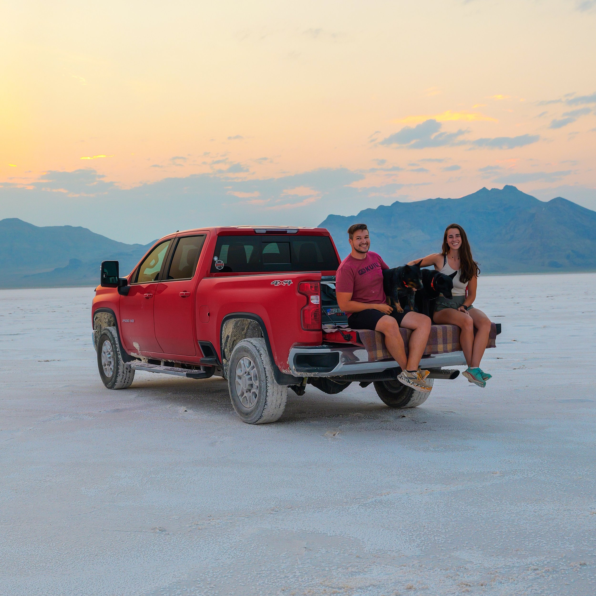 A man and woman sit on the back of a red pickup truck with two dogs, outdoors at sunset, with mountains in the background.