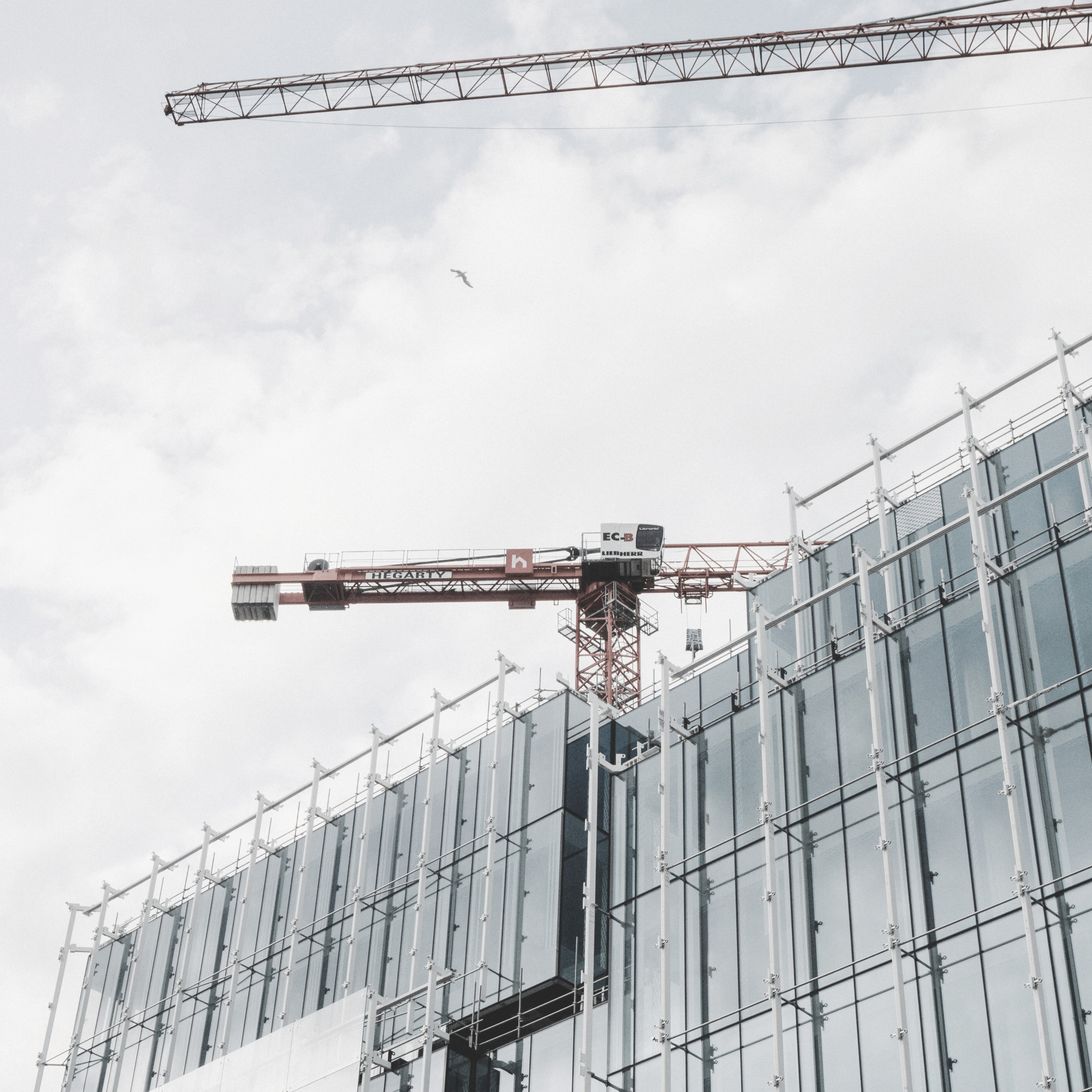 Construction site with glass building and two cranes against cloudy sky, one is red and the other is yellow.