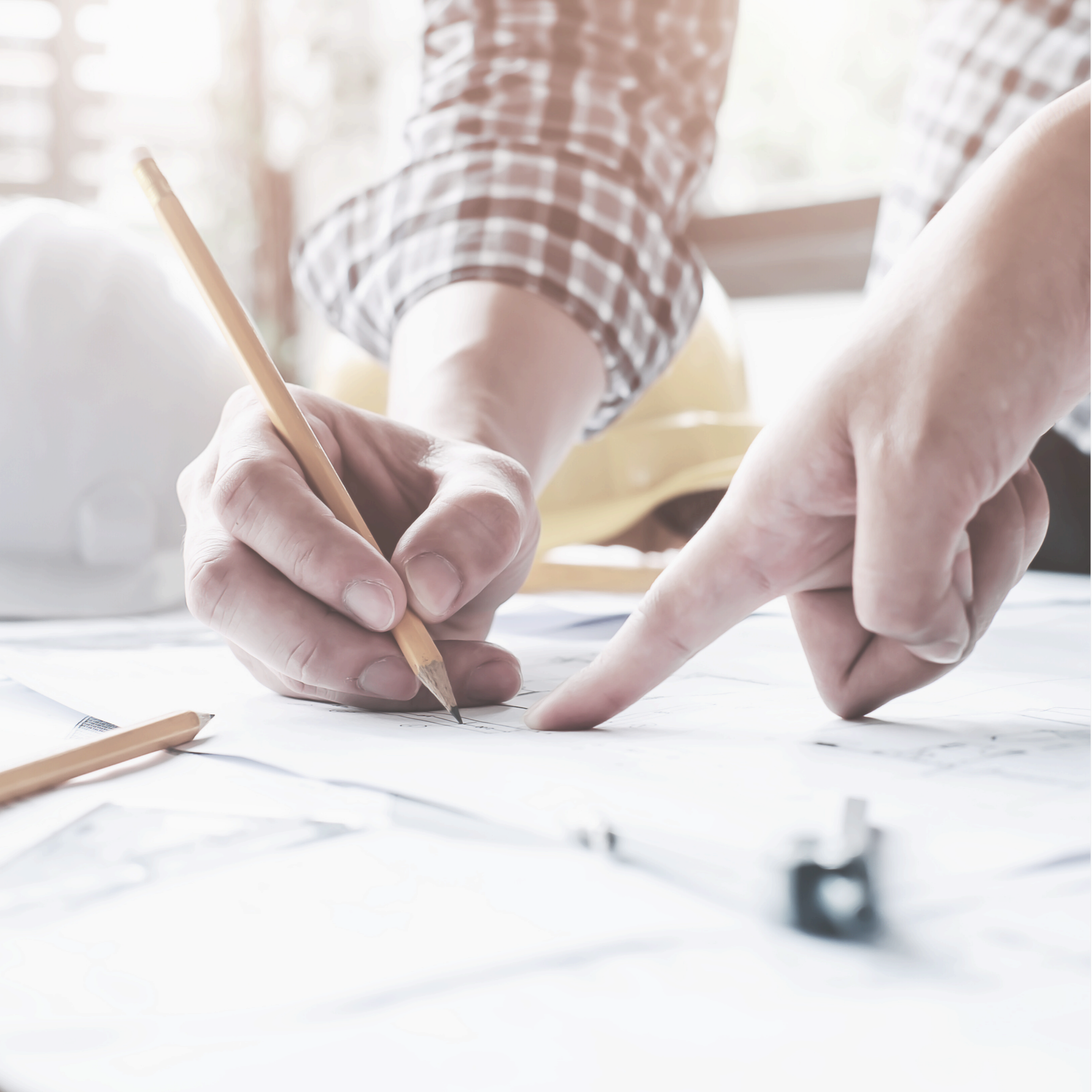 A person in a checkered shirt is drawing or drafting on paper with a pencil, pointing at the paper with their finger.