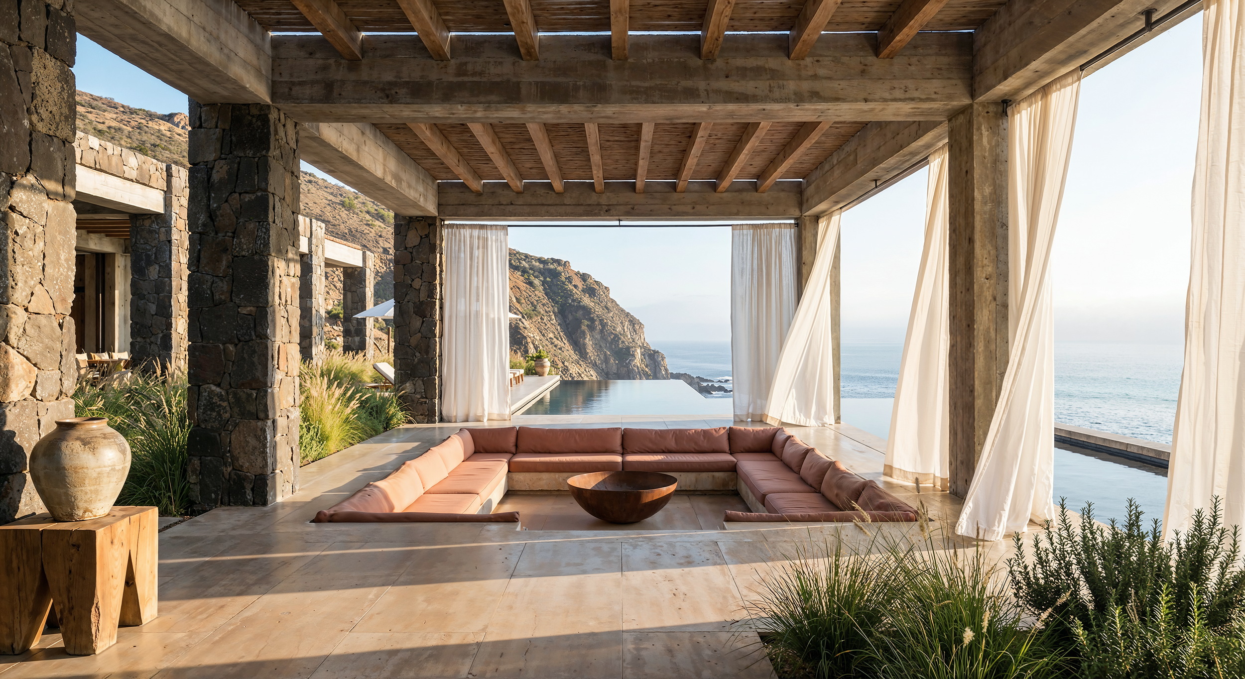 Open-air lounge area with a circular pink sofa and a wooden coffee table, overlooking the ocean with a cliff in the background. The space has beige curtains, stone columns, and a wooden ceiling, surrounded by greenery.
