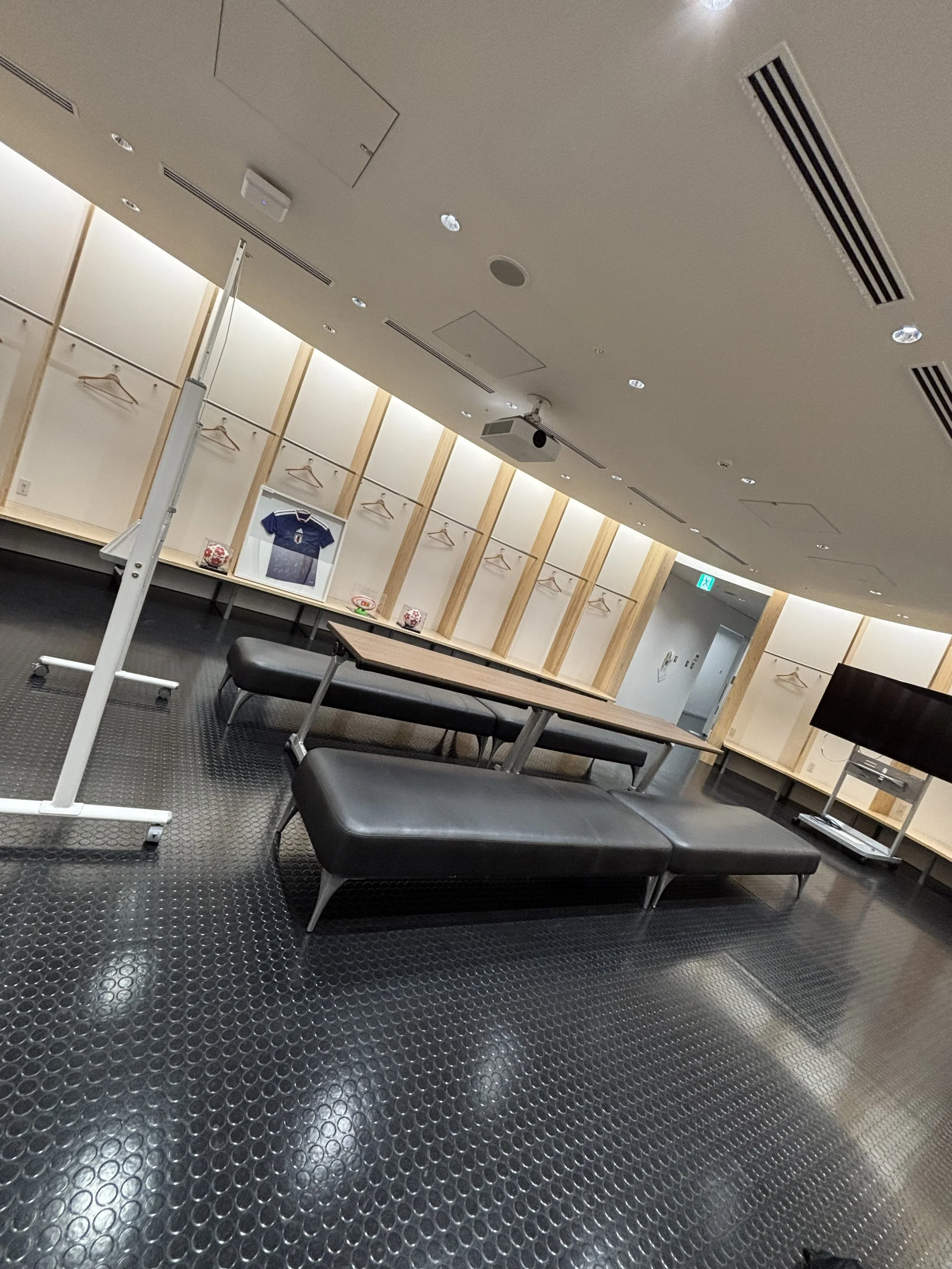 Japan national stadium lockerroom made of light colored wood and has plenty of seating in the middle of the room.