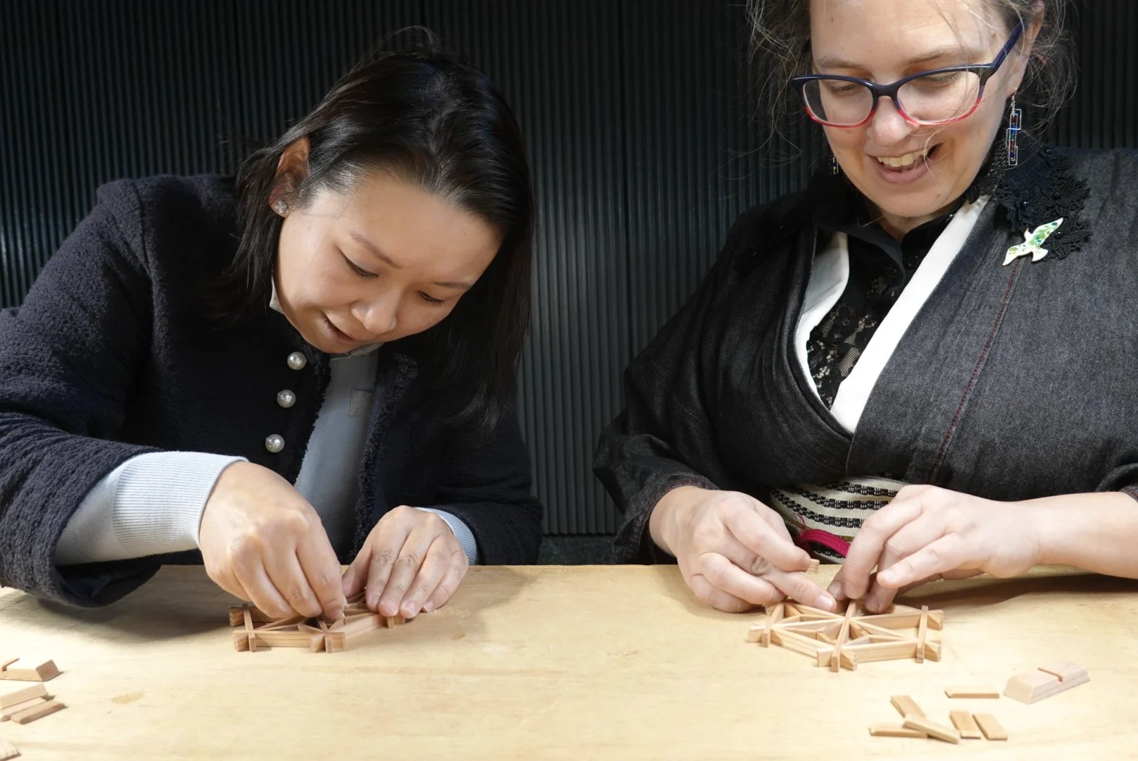 Two woman seated at a table work together to make a Japanese wooden craft held together without glue.