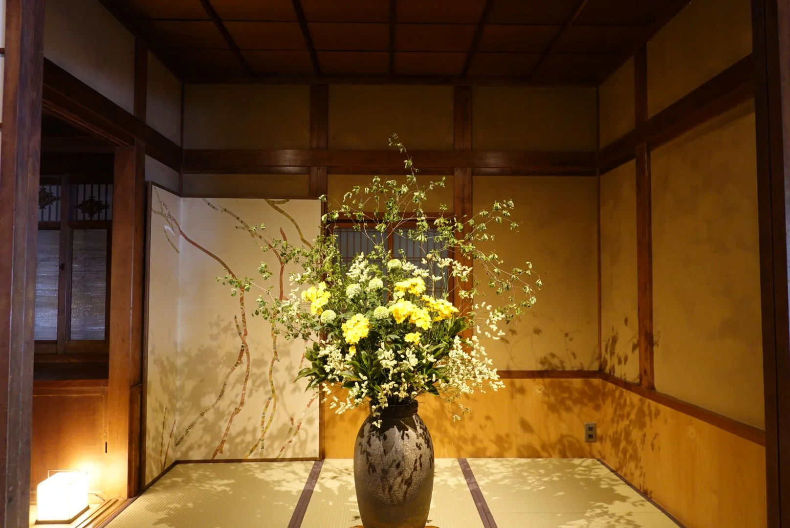 A tall pot of with an ikebana arrangement of yellow flowers and branches stands in the middle of a Japanese tatami mat room with wooden paneling.