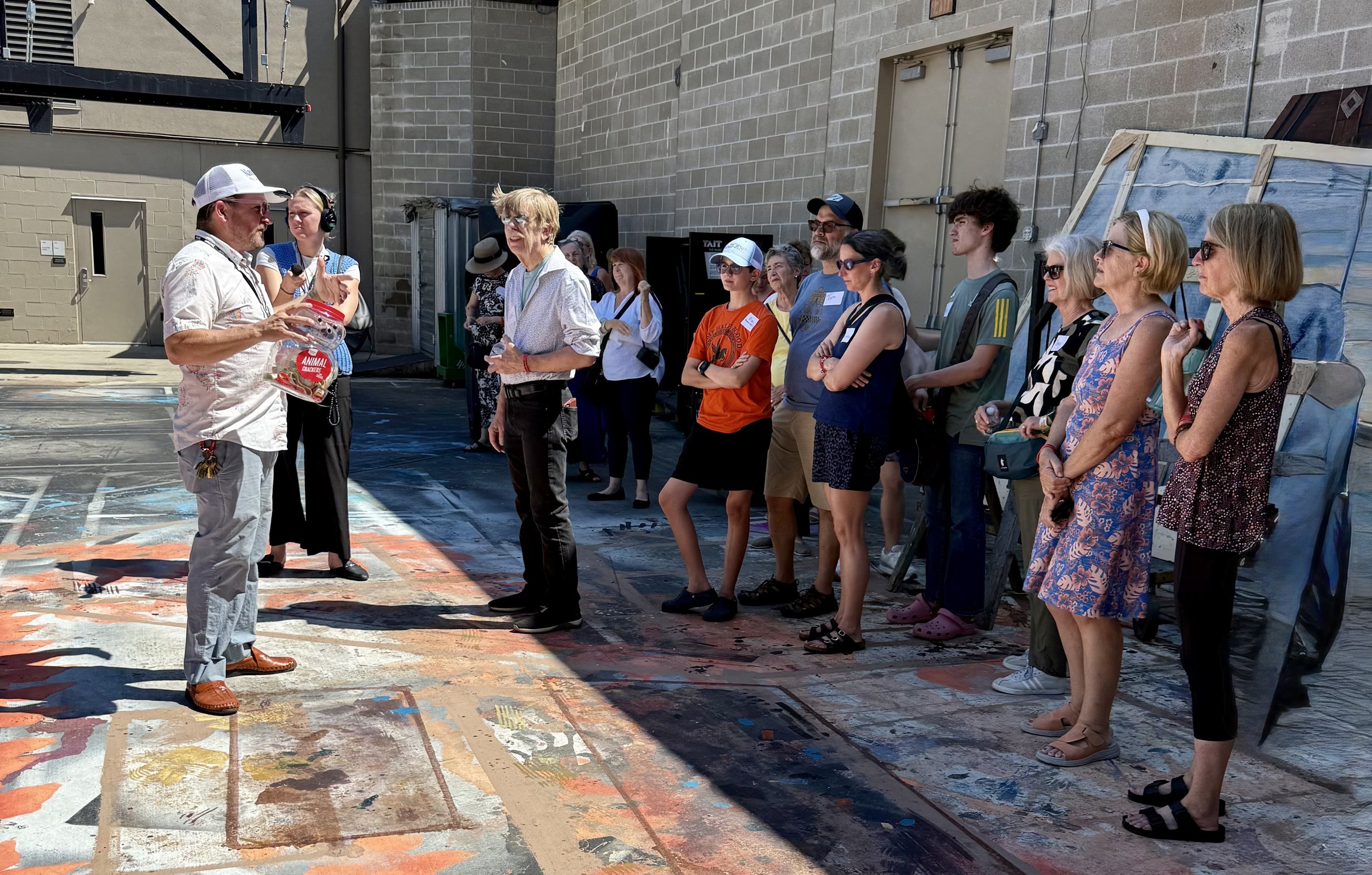 A group of people standing outdoors on a colorful painted ground, listening to a man in a white shirt and hat speaking and holding a plastic bag, while a woman in a white blouse and glasses stands nearby.