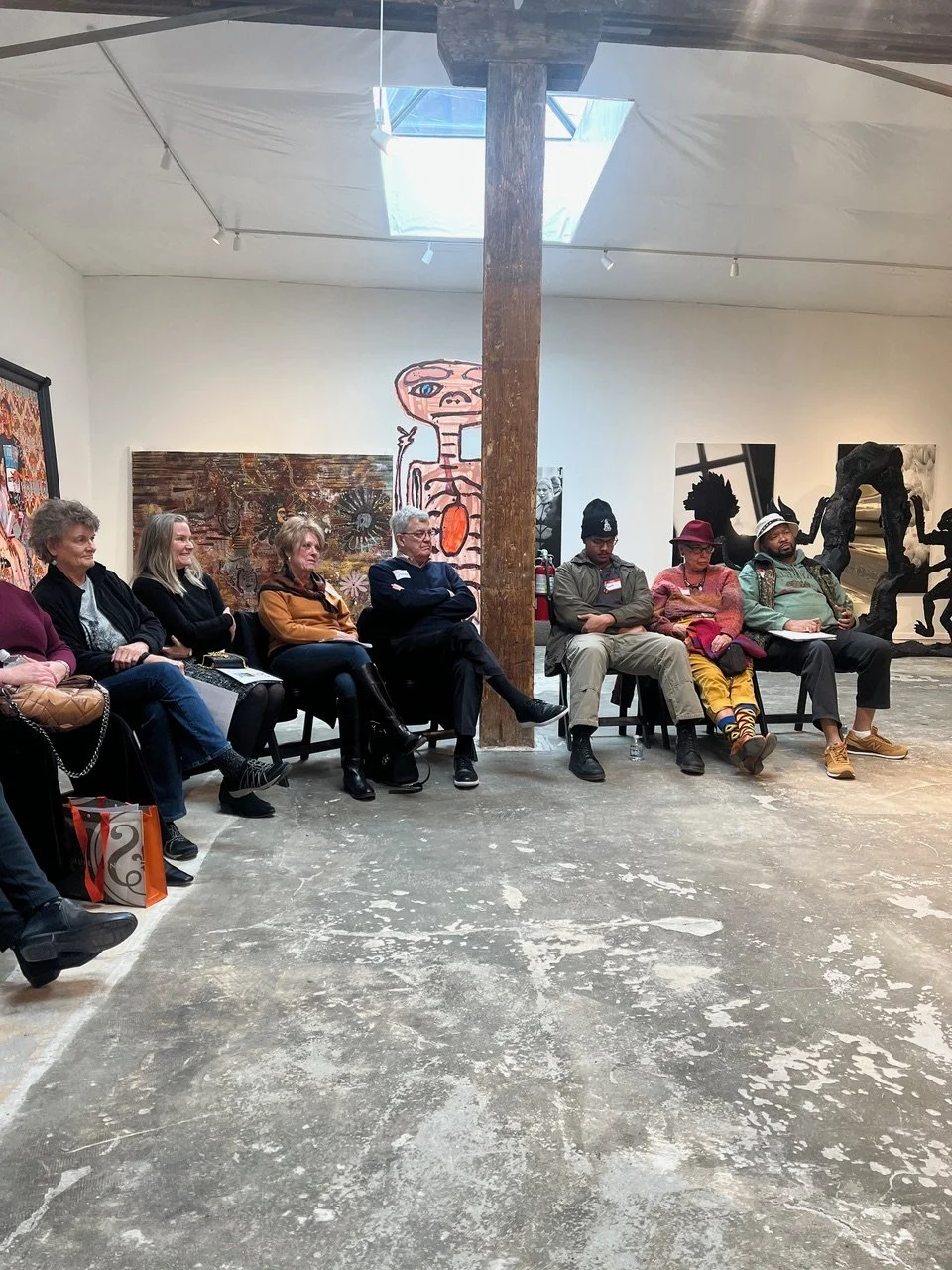 People sitting in a gallery or museum, attending an event or presentation, with artwork displayed on the walls behind them.