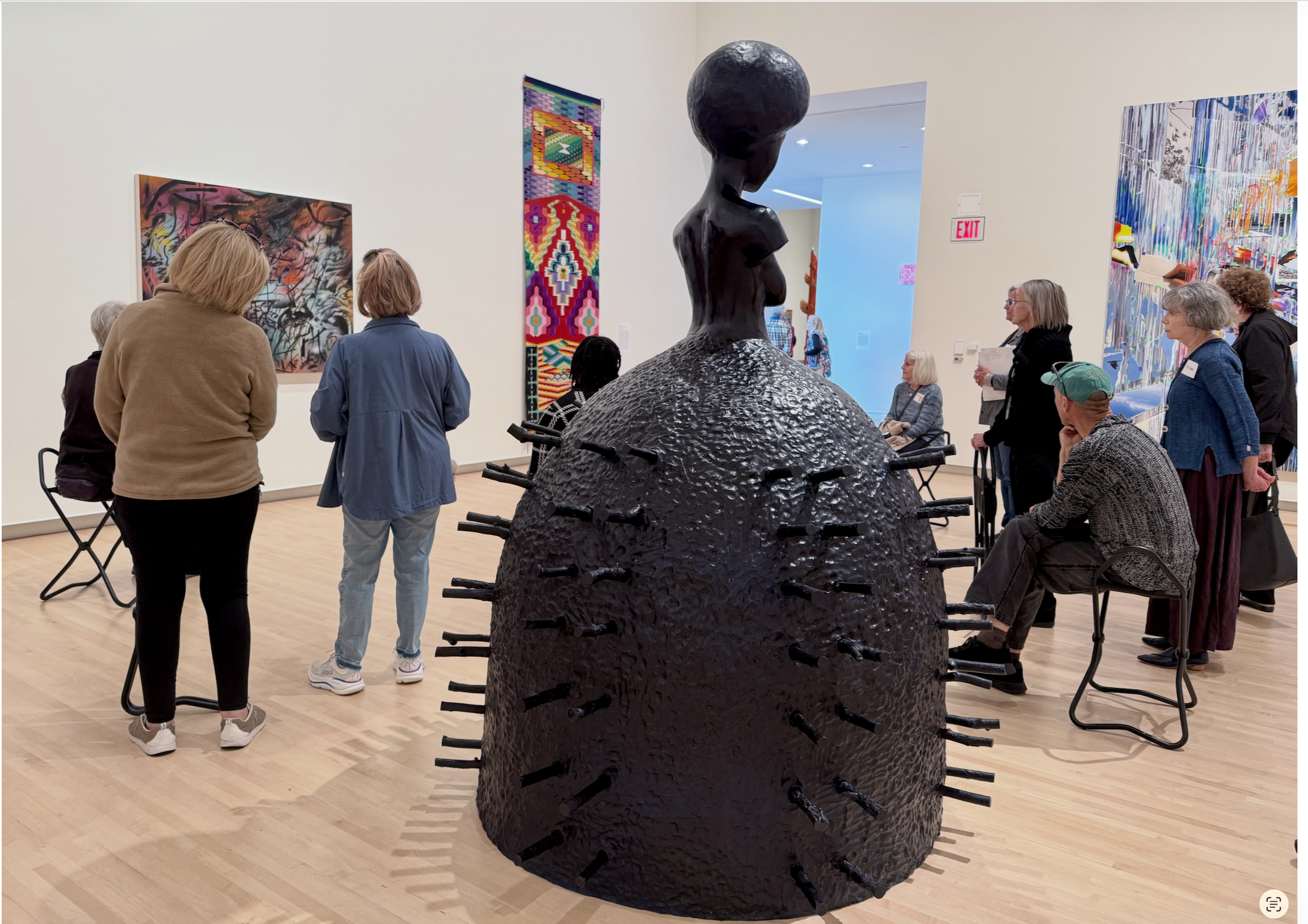 Gallery with a black sculpture of a woman with an oversized head and a textured rounded base surrounded by seated and standing visitors, with colorful abstract paintings on the walls.