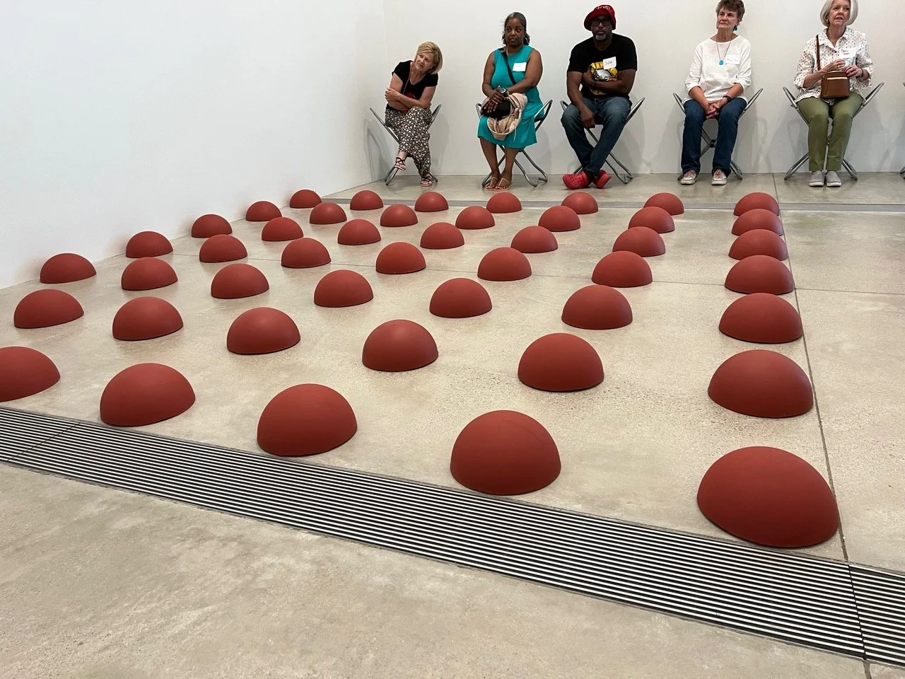 Installation of red semi-spherical seats arranged in grid pattern on beige floor with five people sitting on chairs against white wall in background.