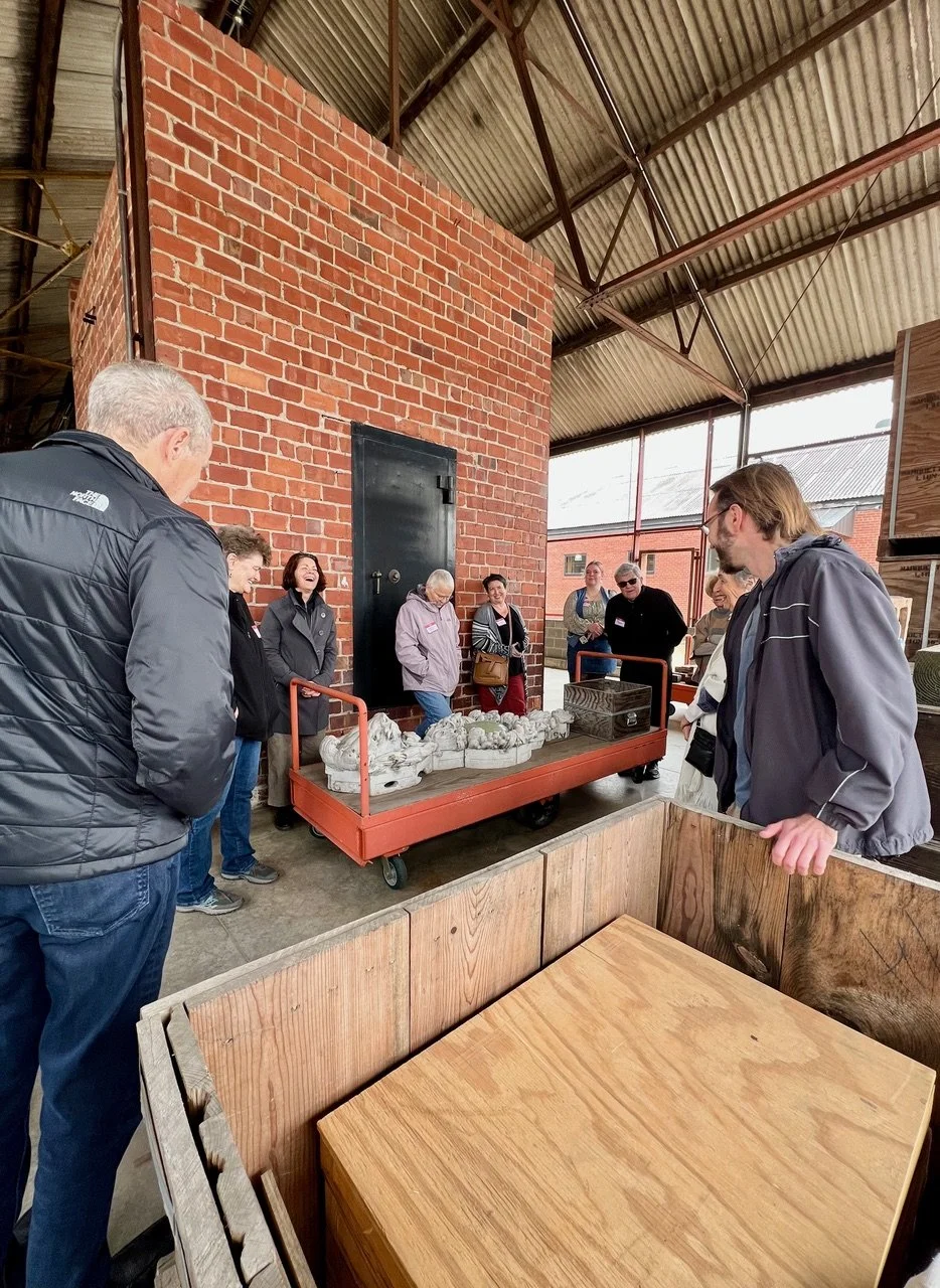Group of people browsing sculptures in a warehouse with brick wall and metal roof