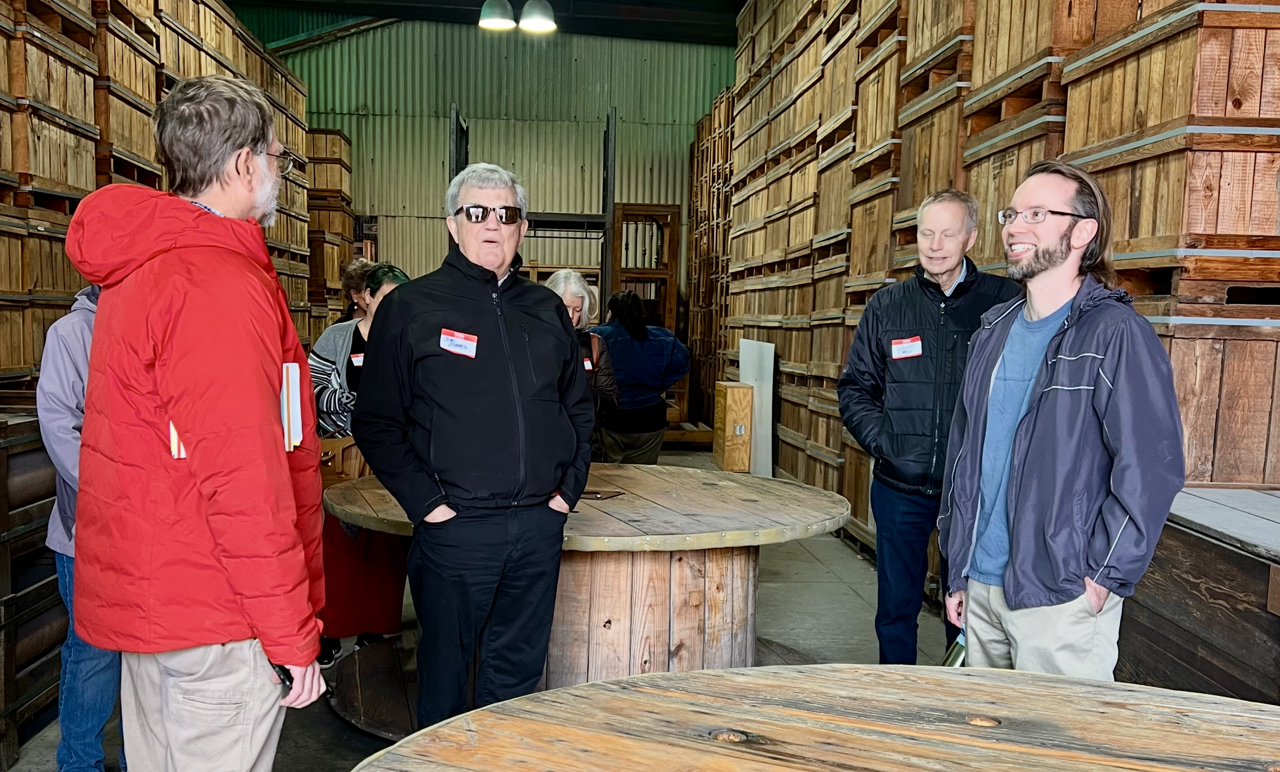 Group of people having a conversation inside a wood warehouse or storage room with large stacks of wooden crates and spools, some wearing name badges and casual jackets.