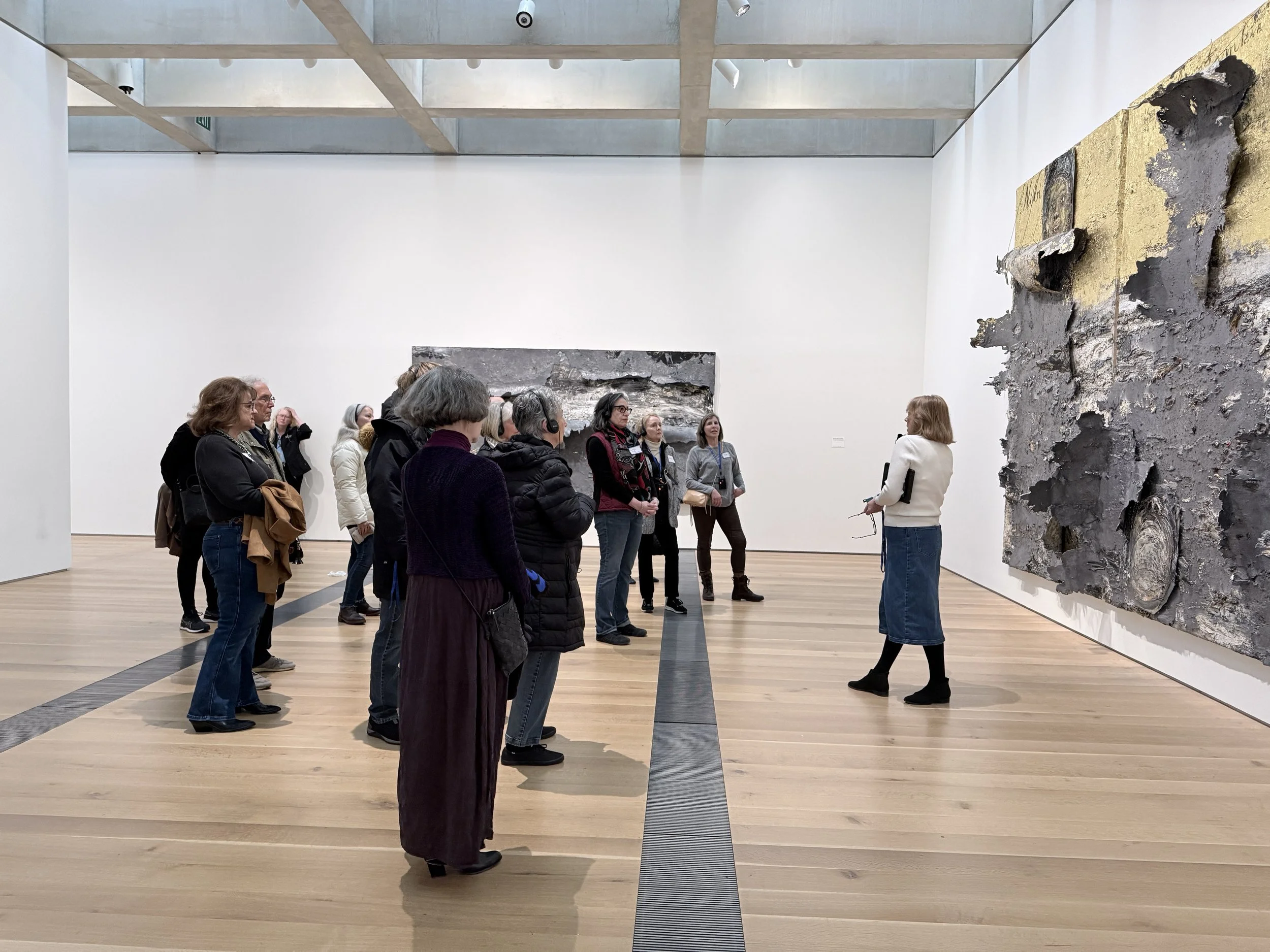 Group of people standing and listening to a woman giving a tour in an art gallery, with large, textured abstract paintings on white walls and light wood flooring.