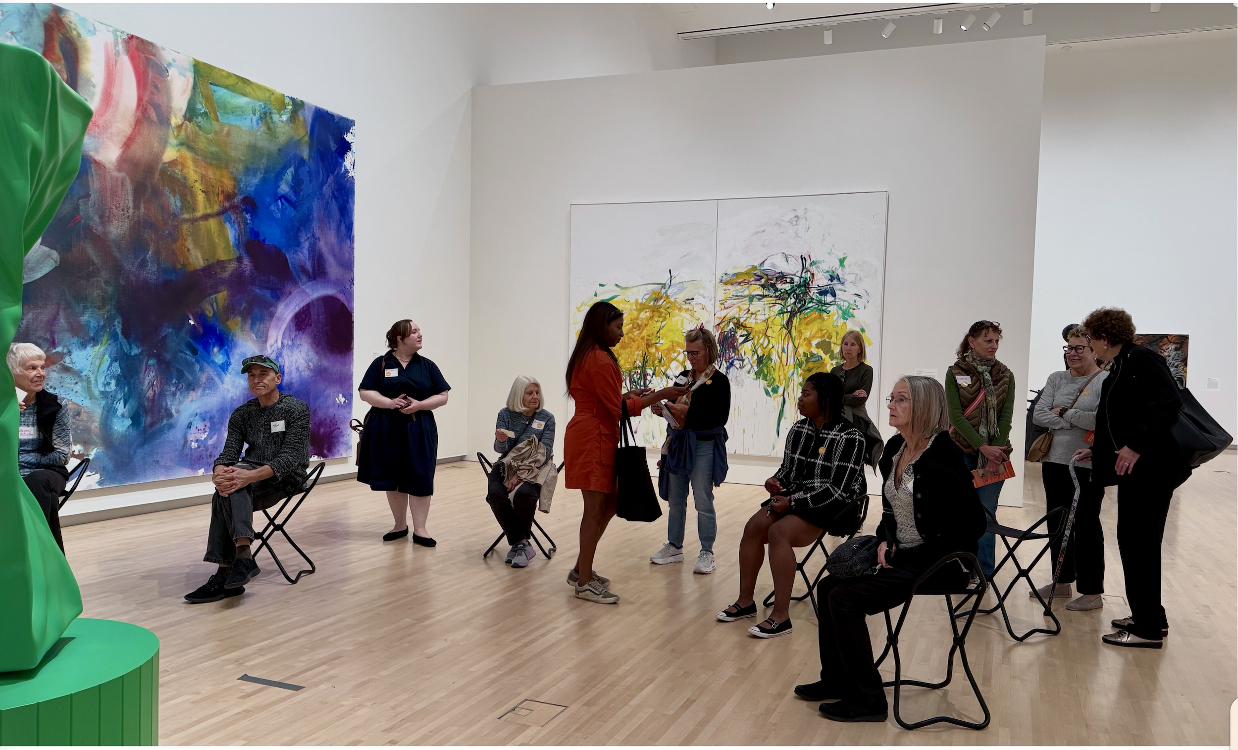 People attending an art gallery exhibit, with abstract paintings on the walls, some sitting and some standing, and one person handing an item to another in the center.
