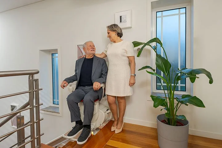 A senior couple in their home, with the husband using a stairlift to navigate the stairs while his wife stands next to him.