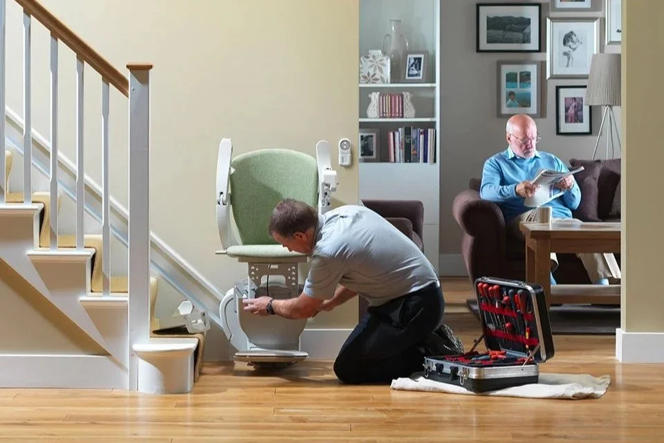 A technician kneels to perform maintenance on the base of a stairlift at the foot of a staircase while an open toolkit sits nearby and a resident relaxes in the background.