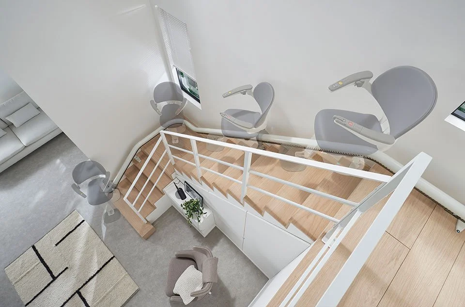 An overhead time-lapse illustration shows a curved stairlift smoothly ascending a light-wood staircase with white railings in a modern, open-plan home.