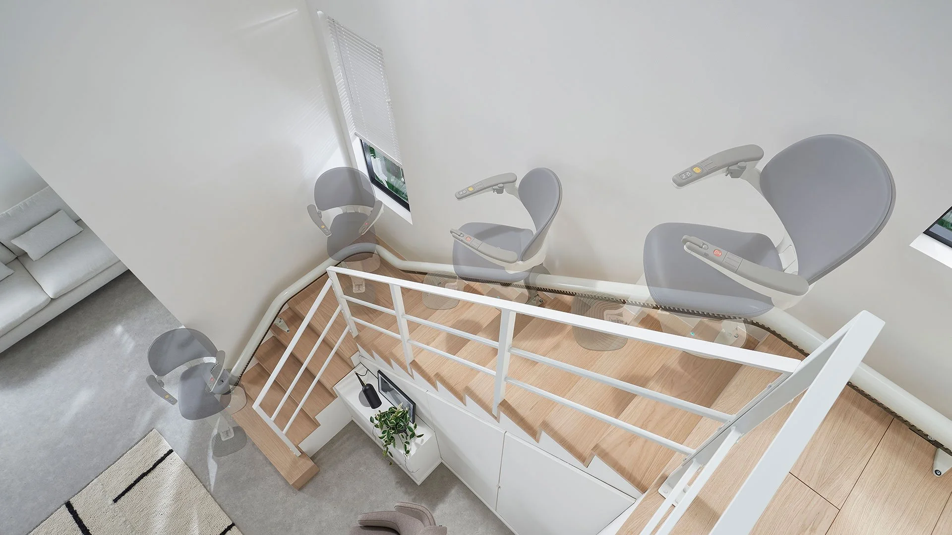 An overhead time-lapse illustration shows a curved stairlift smoothly ascending a light-wood staircase with white railings in a modern, open-plan home.