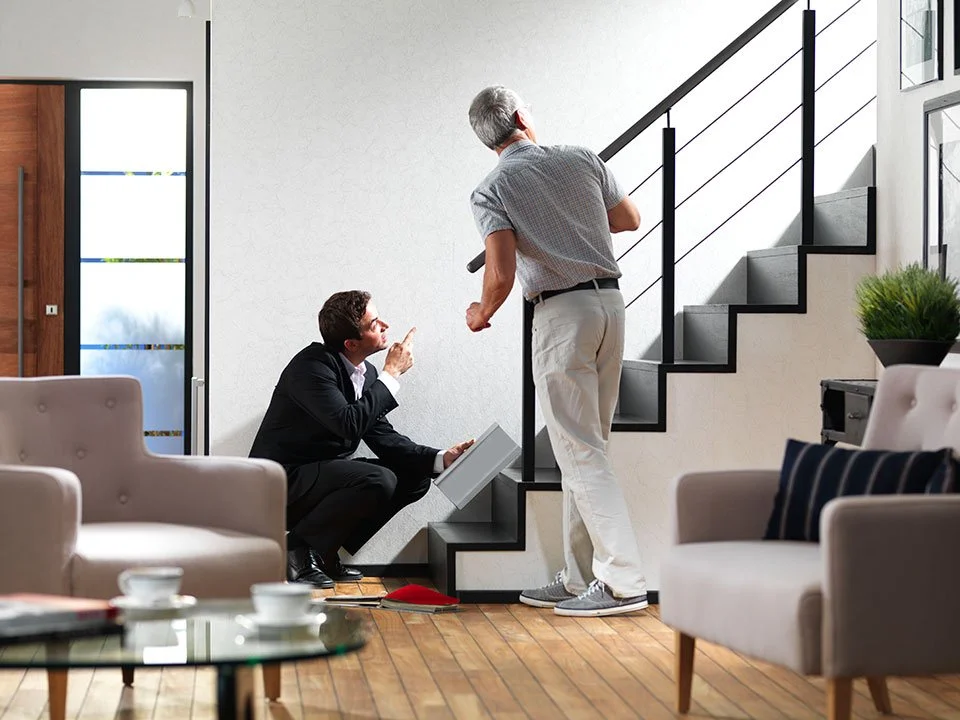A professional consultant in a suit kneels by a staircase, pointing upward and holding a sample while discussing a home accessibility solution with a man standing on the first step.